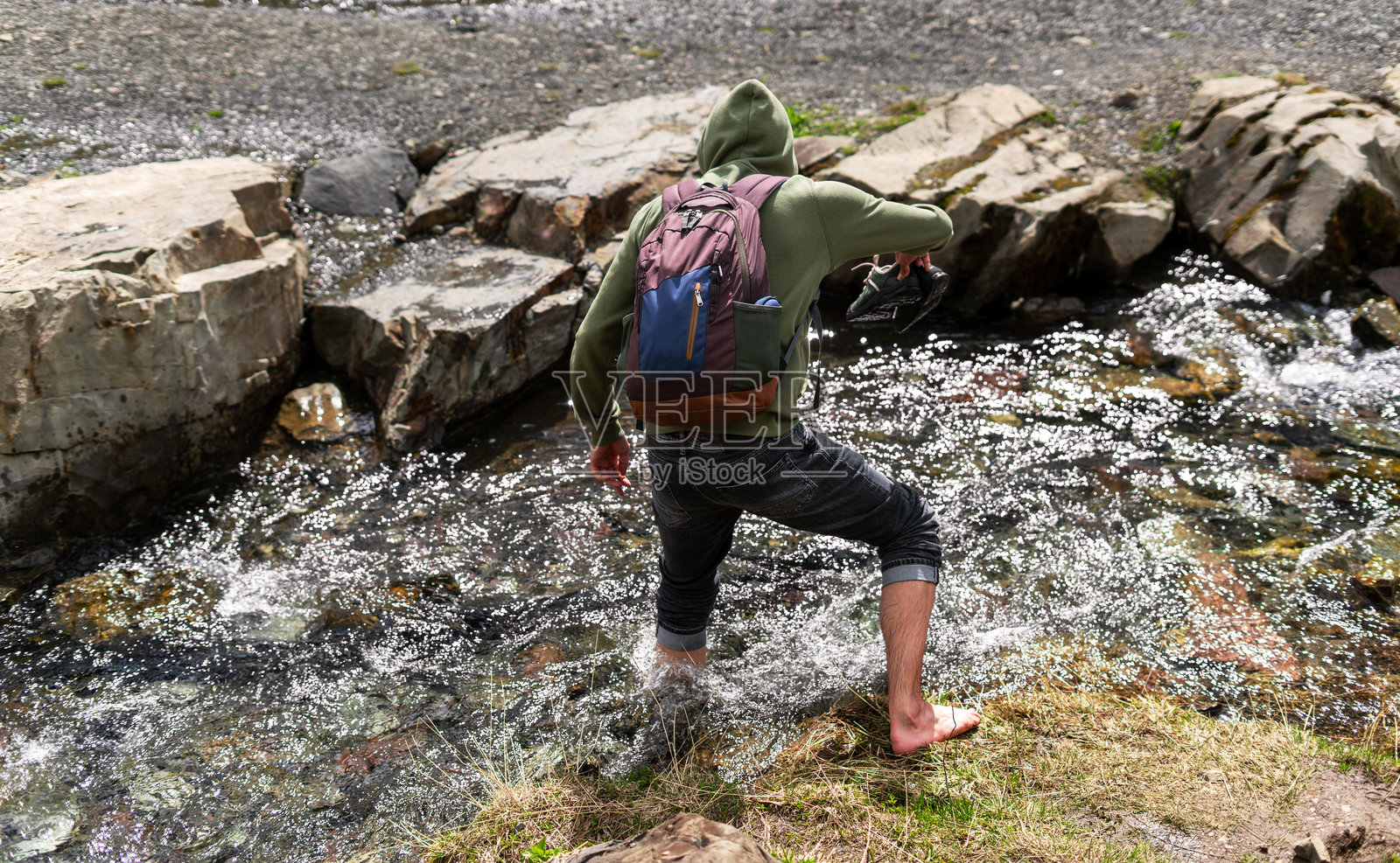 年轻人穿着连帽衫和背包，赤脚穿越岩石小溪，手里拿着鞋子。强调自力更生和平衡的户外冒险与探索，徒步旅行和散步。照片摄影图片