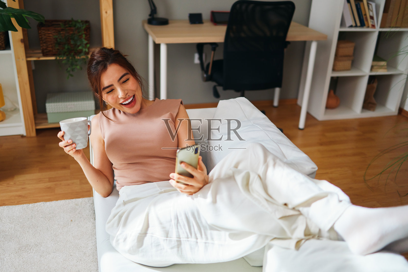 快乐的女人坐在沙发上，使用智能手机并喝咖啡。照片摄影图片