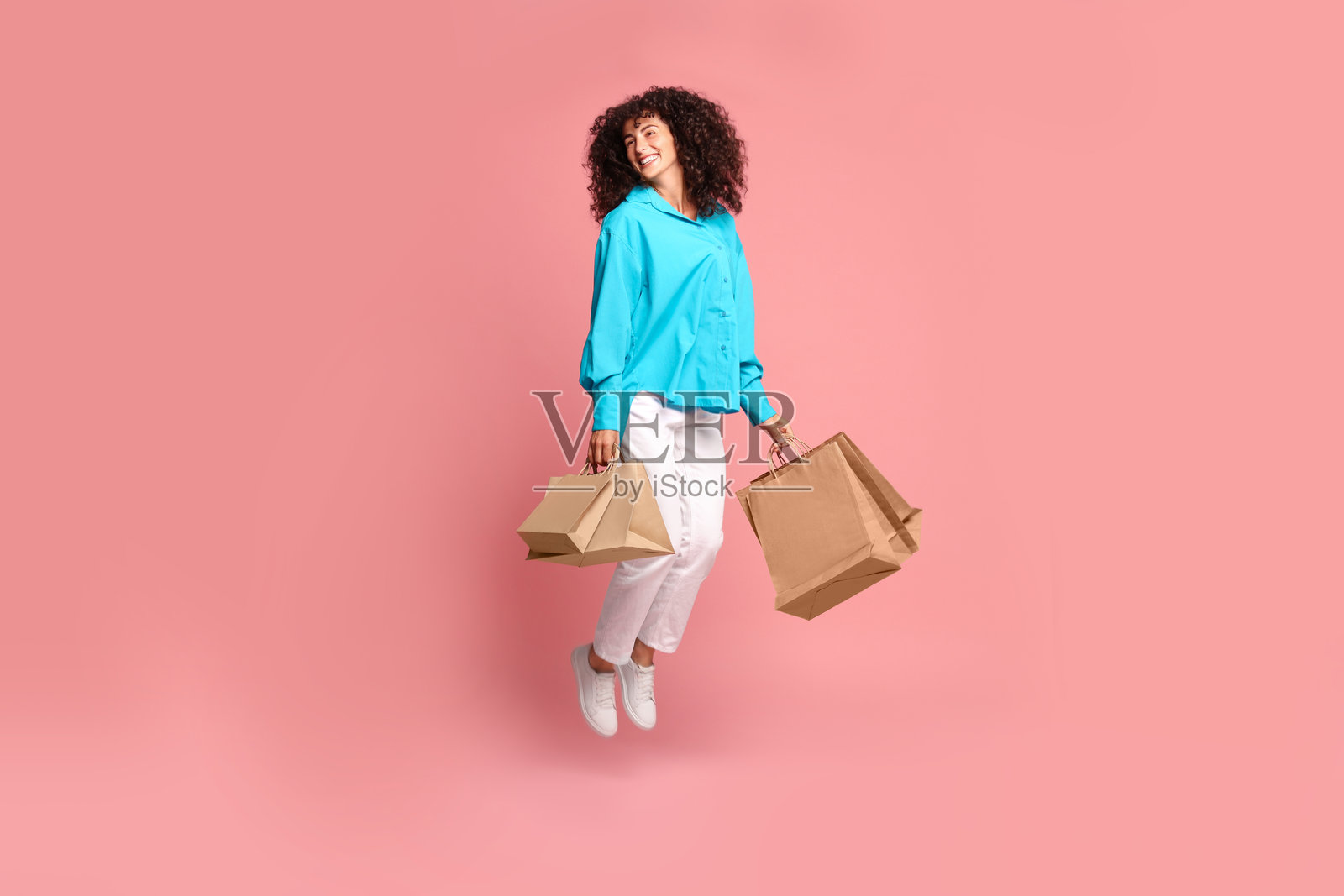 微笑的女性在粉色背景上跳跃，手中提着购物袋照片摄影图片
