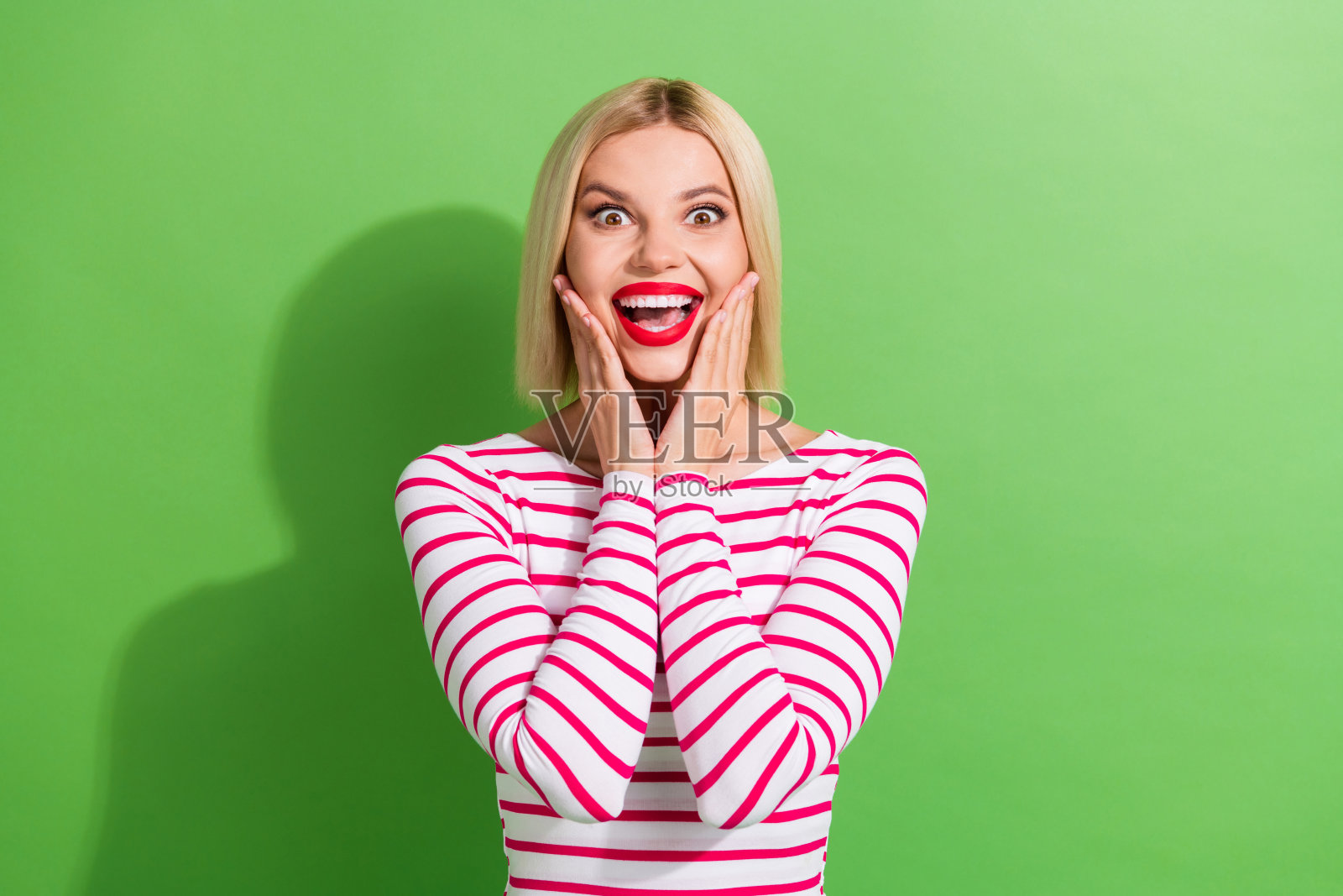 惊讶的短发女孩肖像，穿着时尚的衣服，双手托着脸颊，目不转睛地盯着绿色背景。照片摄影图片