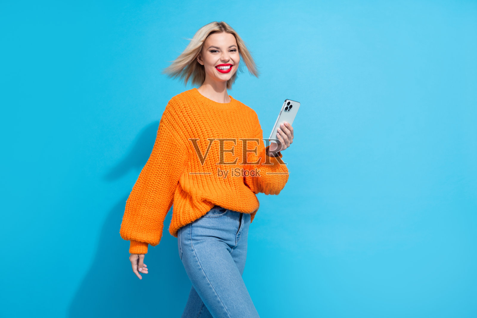 聪明可爱的金发女孩穿着橙色针织毛衣，手持智能手机在蓝色背景下阅读电子邮件。照片摄影图片