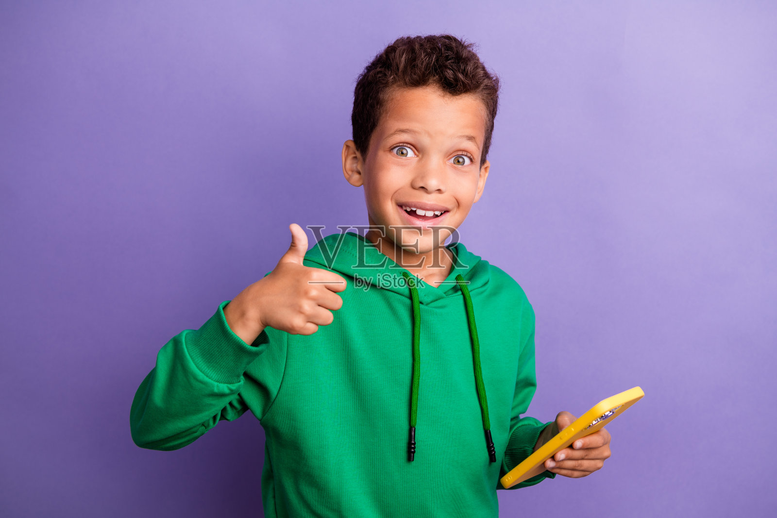 可爱的自信小男孩穿着绿色连帽衫，正在用现代设备发短信，竖起大拇指，背景为紫色。照片摄影图片