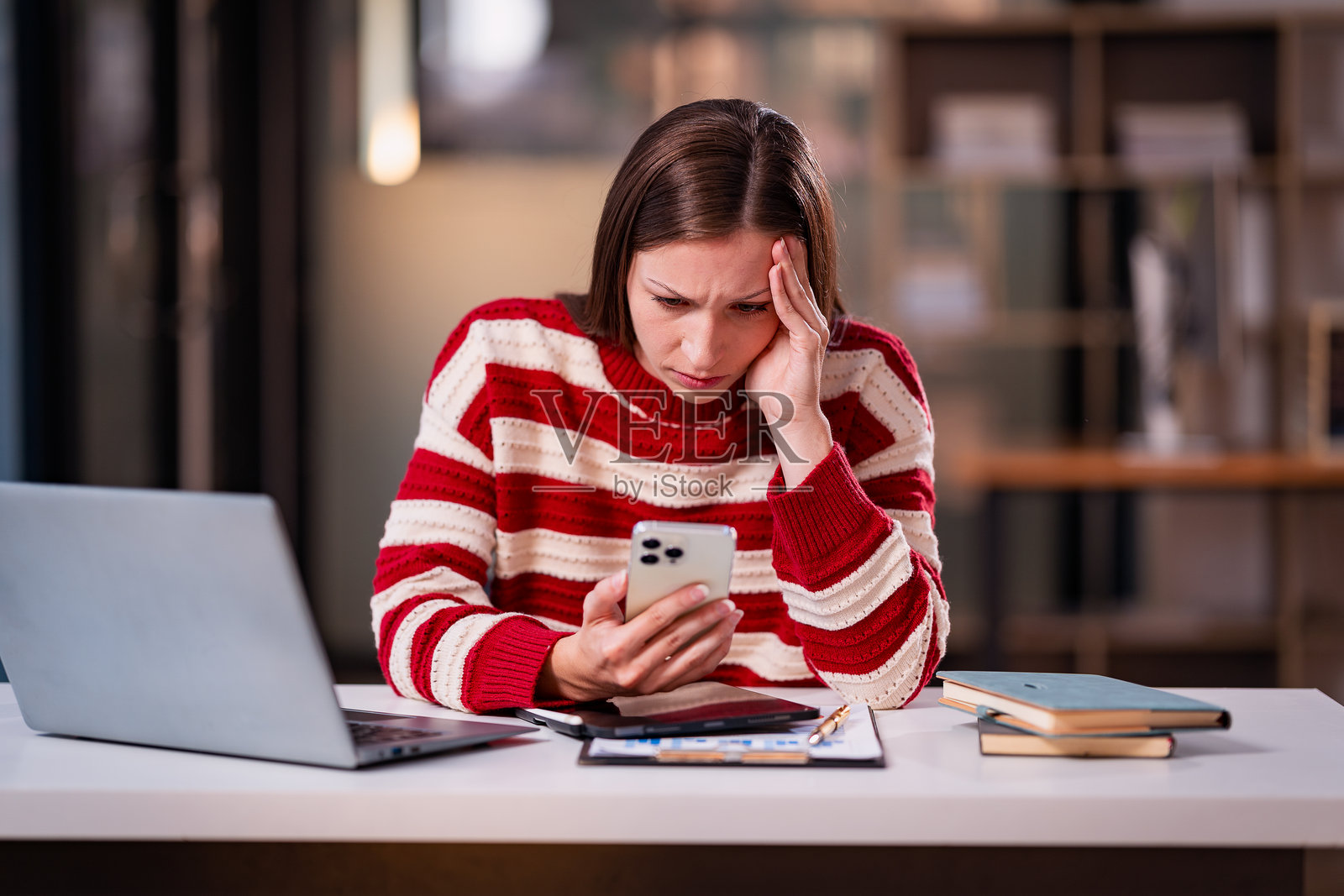 担忧的年轻女性坐在桌前，盯着智能手机屏幕，抚摸着头，表情痛苦，似乎在经历压力或接到坏消息。照片摄影图片