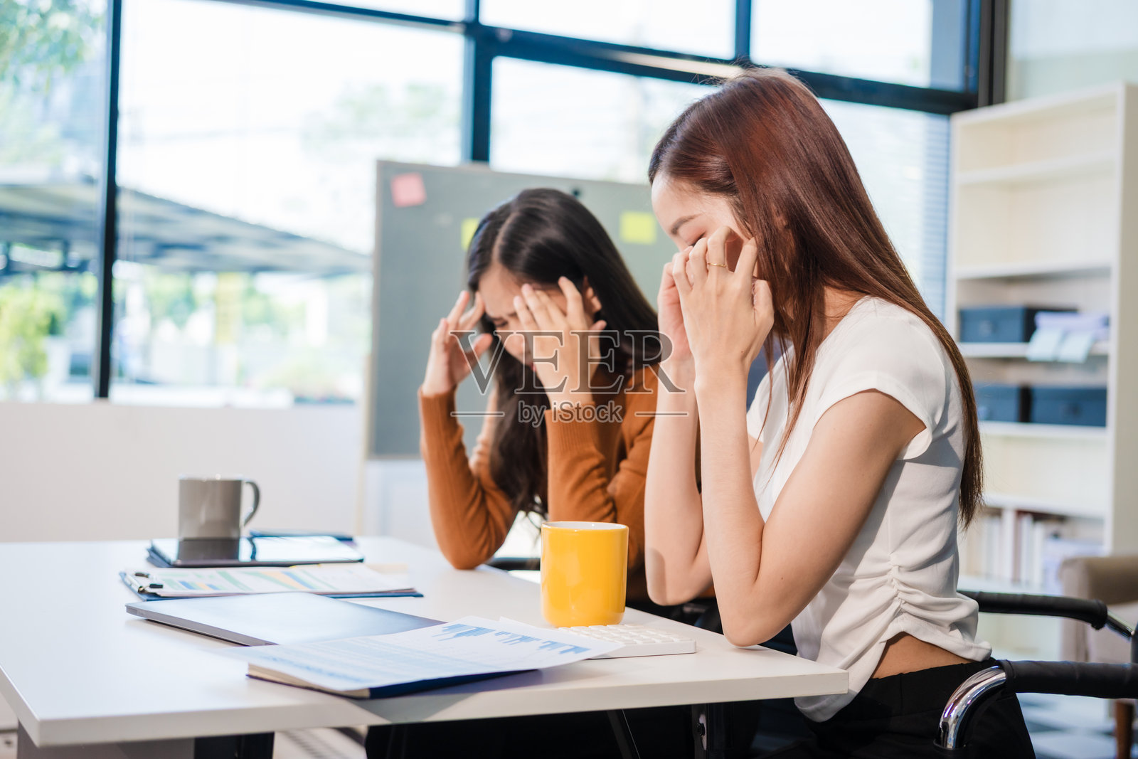 两位年轻的亚洲女商人在办公室桌前坐着。一位女性看起来很紧张，捂着头，而另一位则认真地观察着。她们正面临一个具有挑战性的工作情况，气氛紧张。照片摄影图片