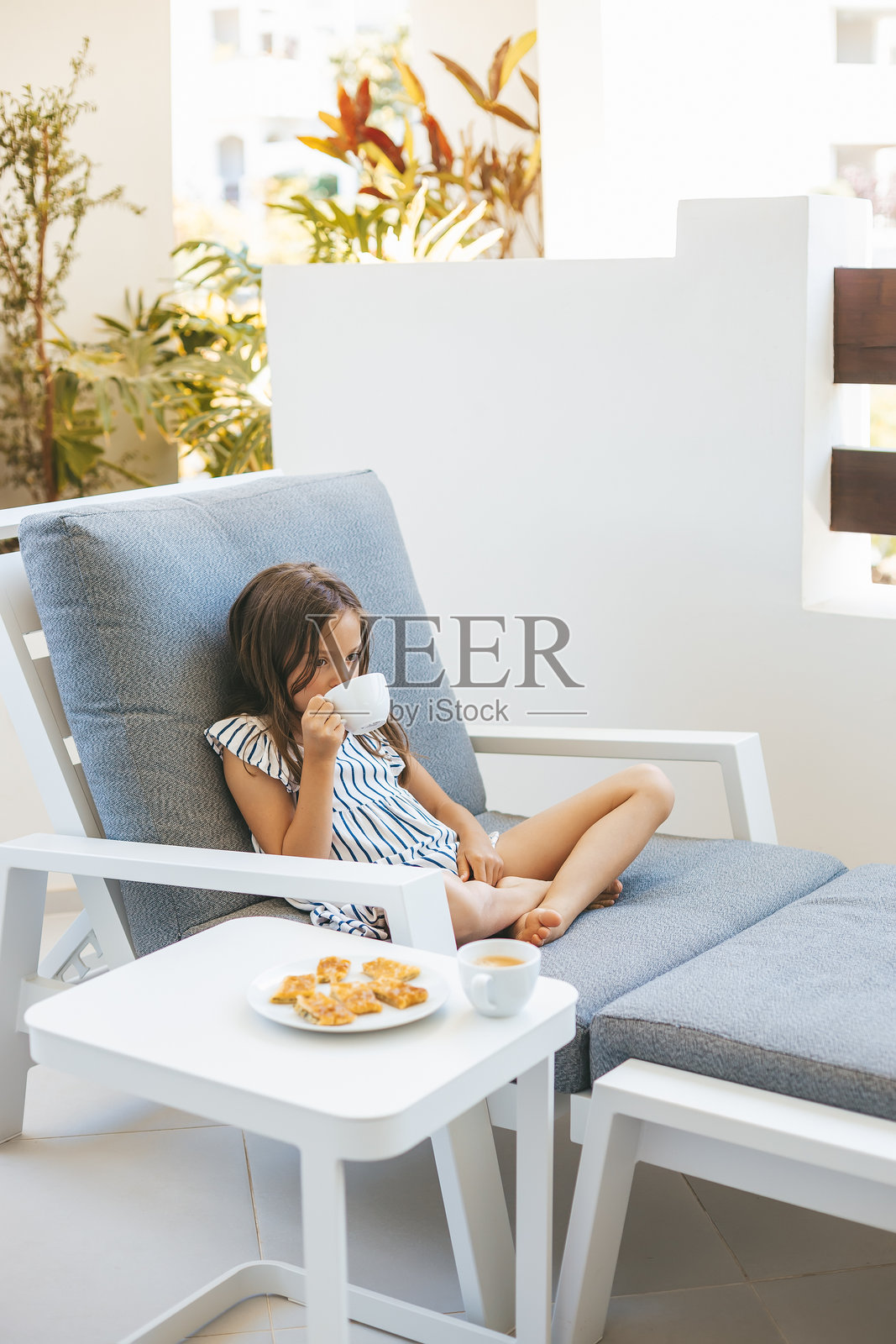 穿着条纹裙的小女孩在露台躺椅上放松，手里端着茶，神情若有所思。照片摄影图片