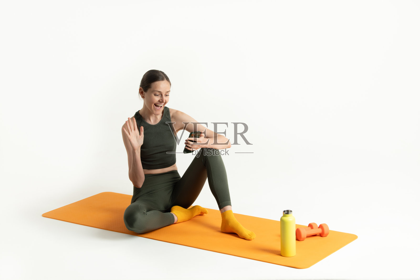 一位穿着运动服的女性坐在橙色瑜伽垫上，微笑着使用手机。旁边有小哑铃和水瓶，突显了她的健身日常。照片摄影图片