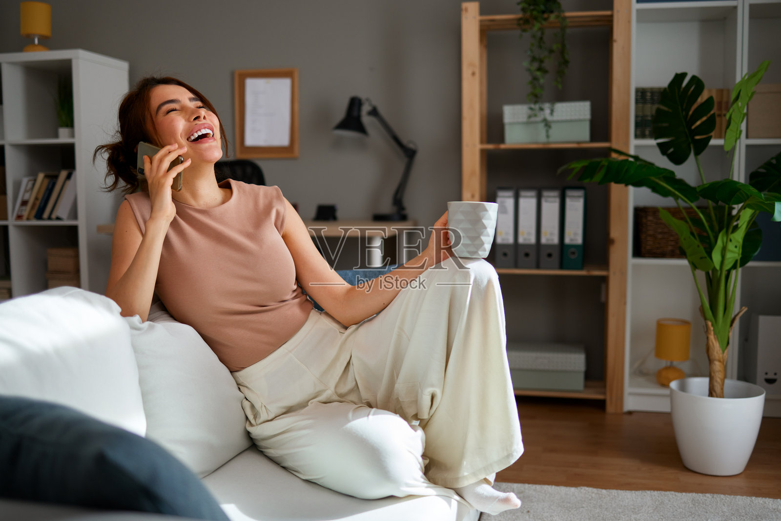 快乐的年轻女性在家沙发上放松，喝着咖啡并打电话。照片摄影图片