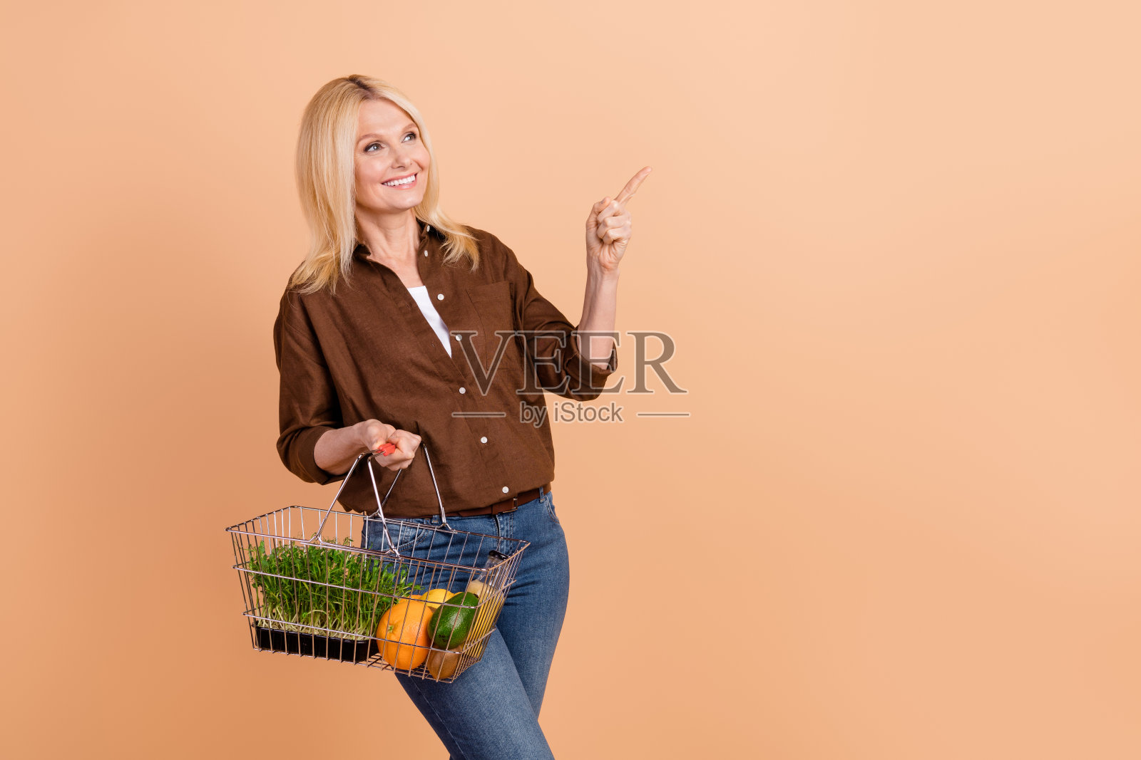 穿着时尚棕色衬衫的金发素食女性用手指指向空白处，旁边有装满水果的购物车，背景为米色。照片摄影图片