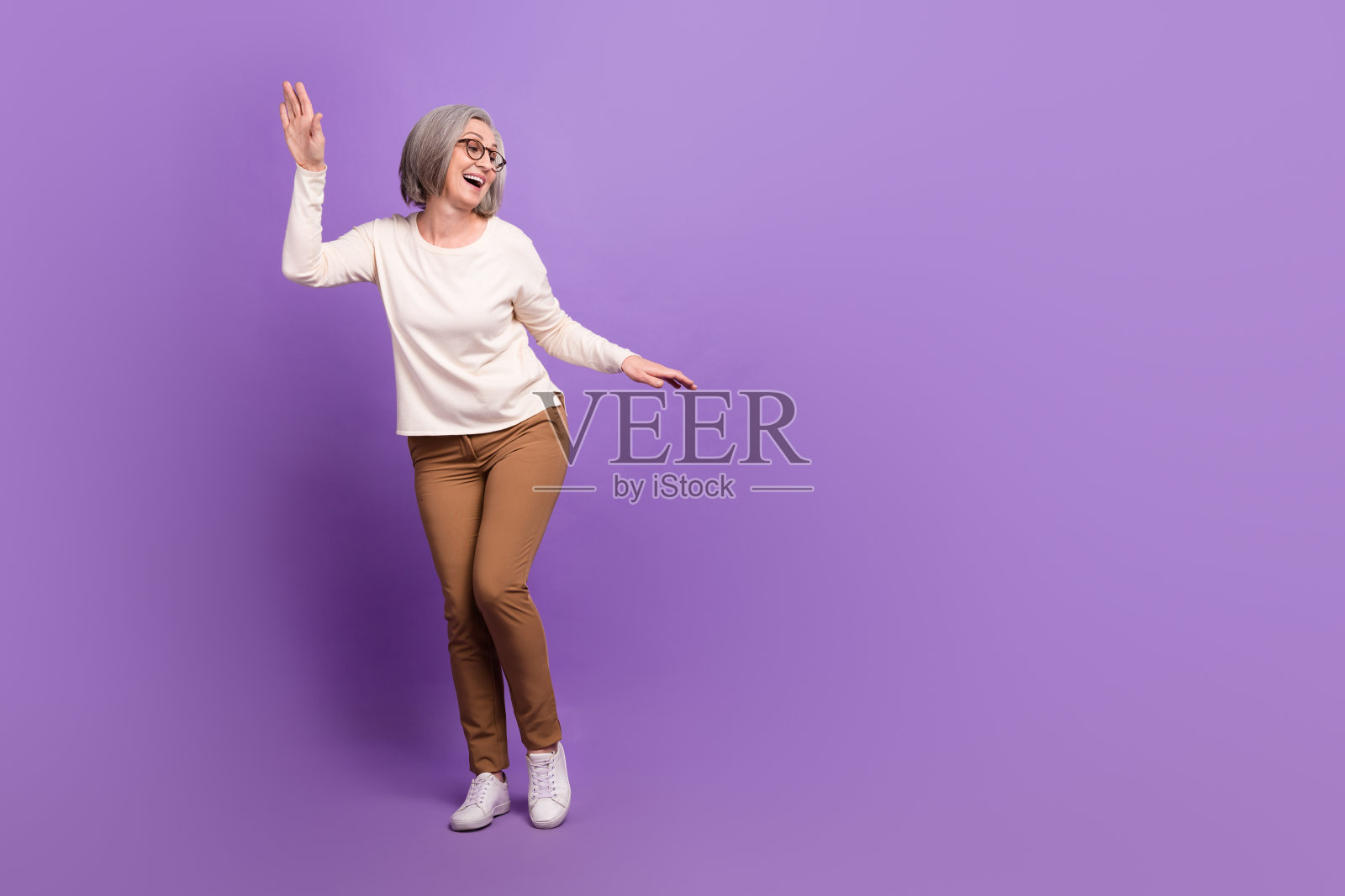 迷人的老年女士全身肖像，跳舞展现年轻派对氛围，穿着时尚休闲服，背景为紫色。照片摄影图片