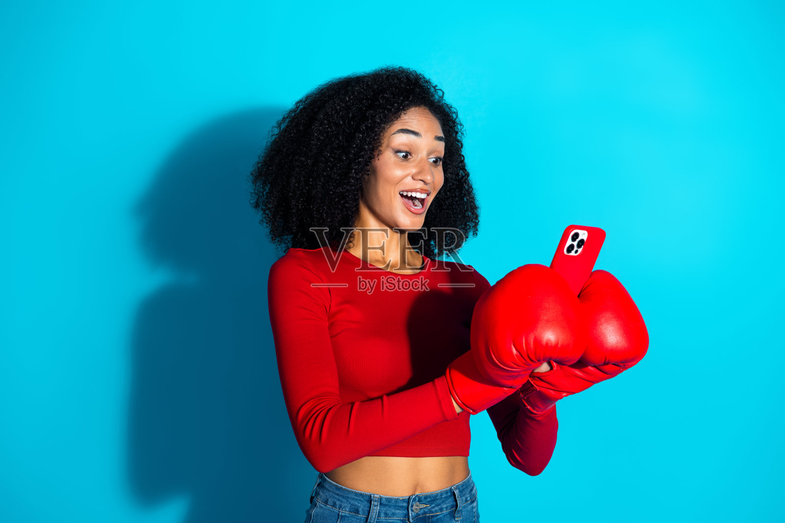 兴奋的年轻女性戴着拳击手套，手持智能手机，背景为蓝色，体验着快乐的惊喜。照片摄影图片