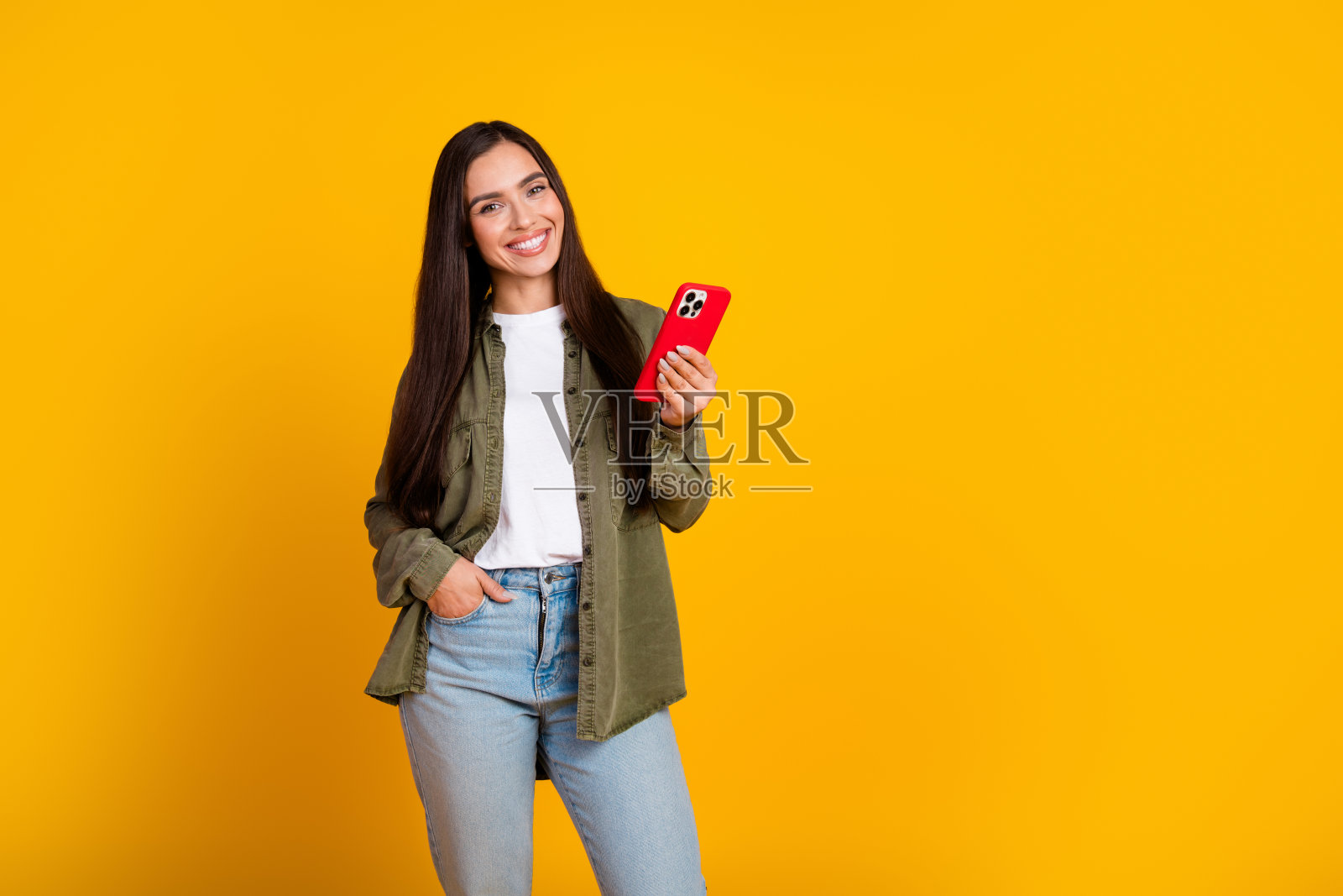 快乐的棕发女性穿着休闲服，手持智能手机，背景为鲜艳的黄色。照片摄影图片