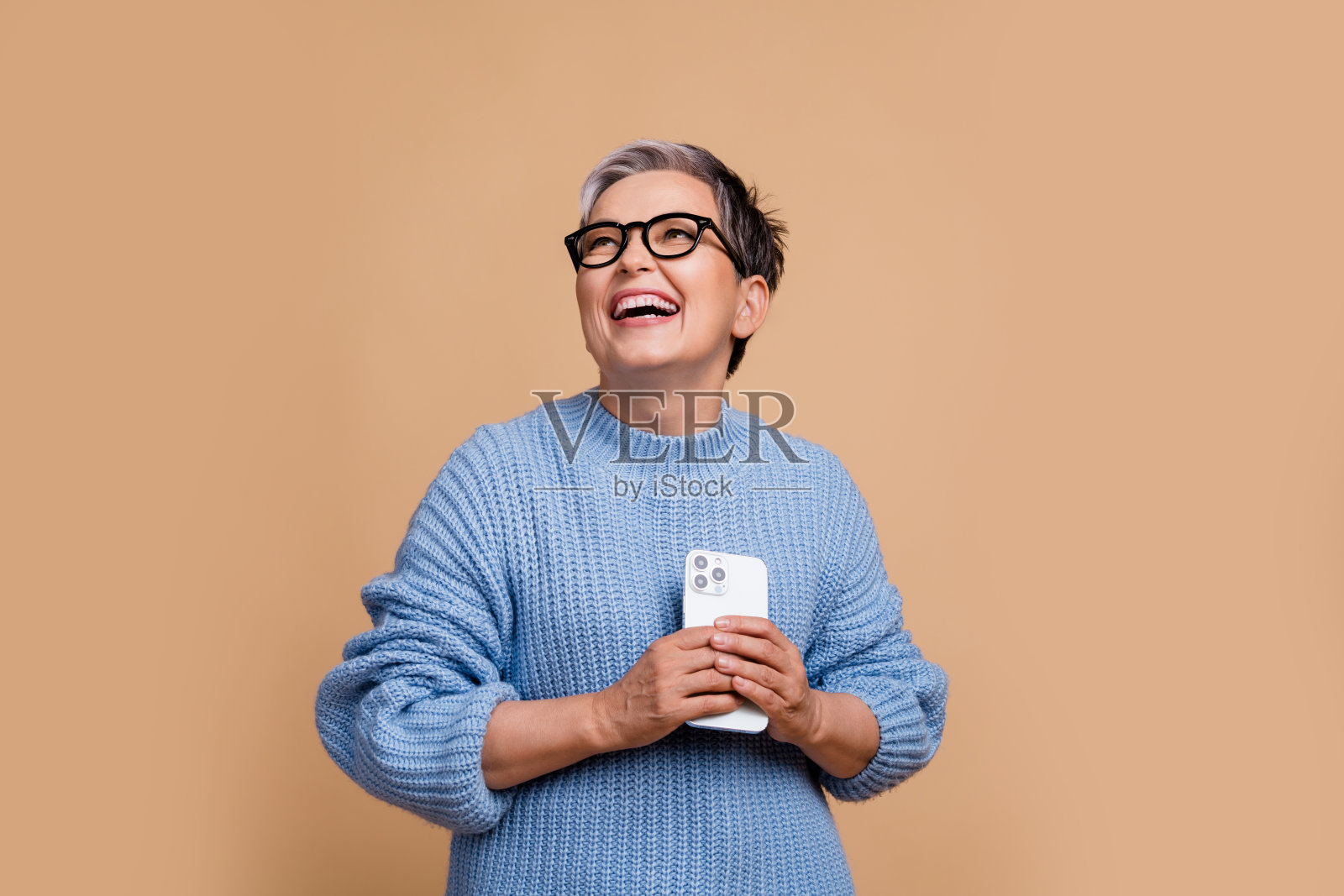 积极的时尚奶奶露出灿烂的笑容，手持智能手机，目光投向空旷的空间，背景为米色。照片摄影图片