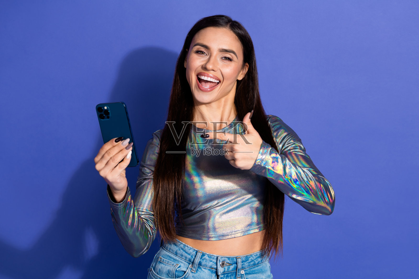 穿着全息服装的快乐年轻女性在紫色背景下指着智能手机，展示时尚潮流与现代科技。照片摄影图片