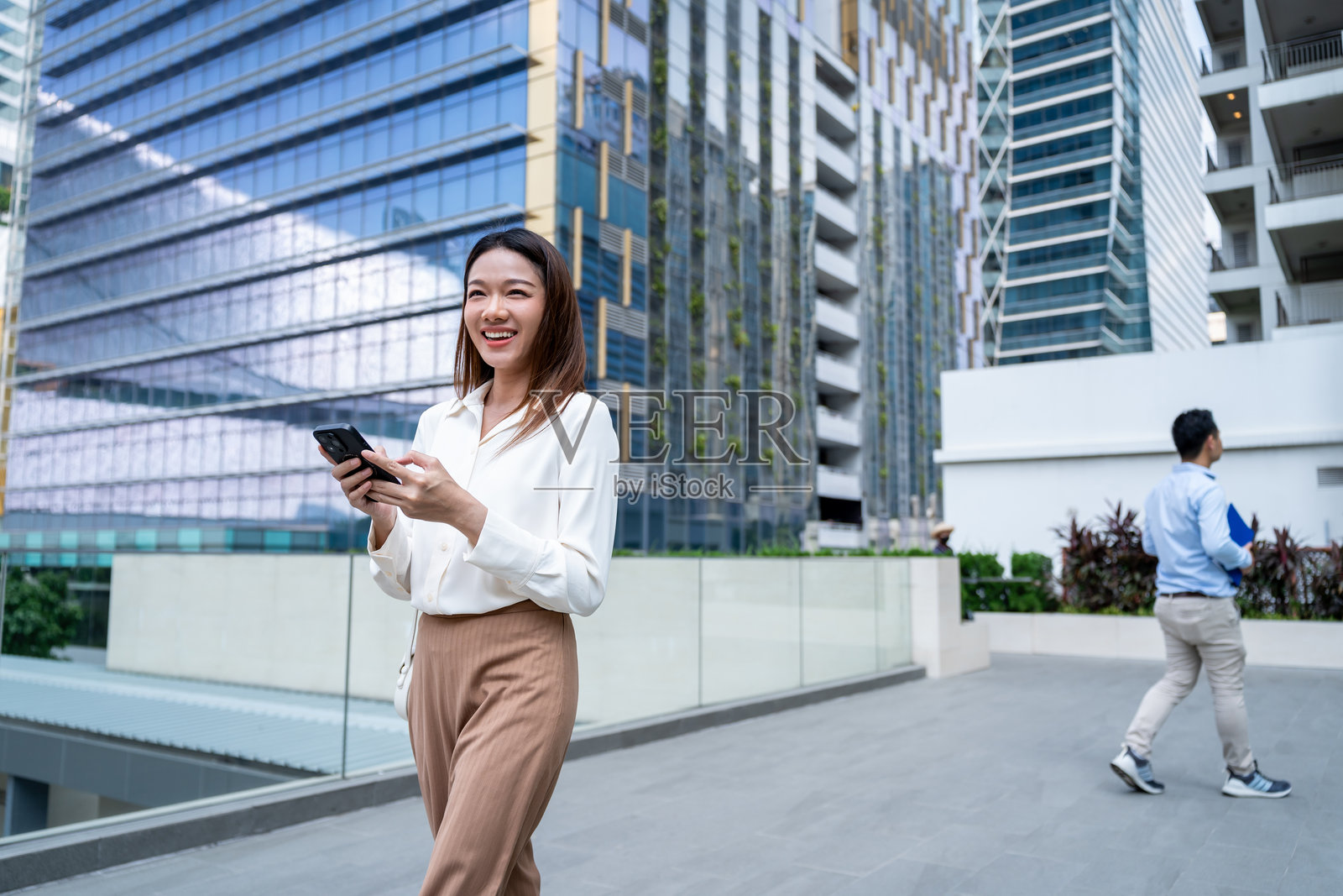 亚洲年轻女性手持手机，浏览社交媒体内容。迷人的美丽女商人员工在现代城市中走动，户外高楼大厦间使用智能手机聊天。照片摄影图片