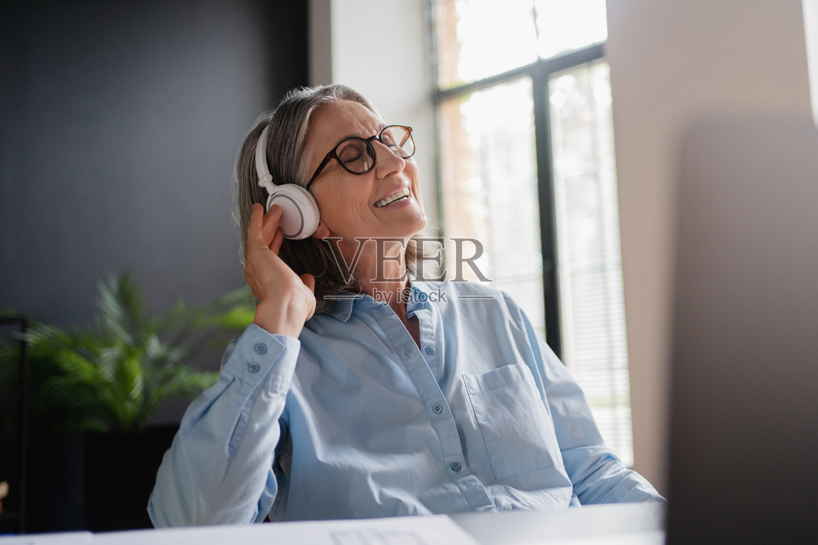 在办公室跳舞的女性，手持笔记本电脑和耳机，面带微笑，享受音频流媒体订阅。舞蹈经理、员工和成熟人士在工作场所使用电脑，在线听音乐。照片摄影图片