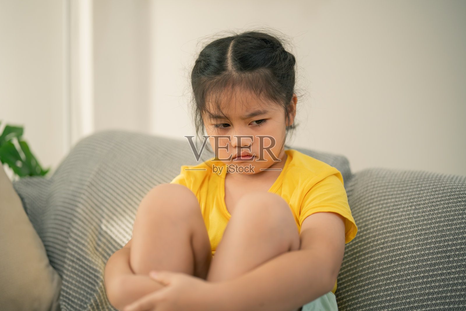 一个坐在沙发上、低头思考的伤心孩子，穿着黄色衬衫，表情沉思，显得孤独，周围是明亮的房间。照片摄影图片