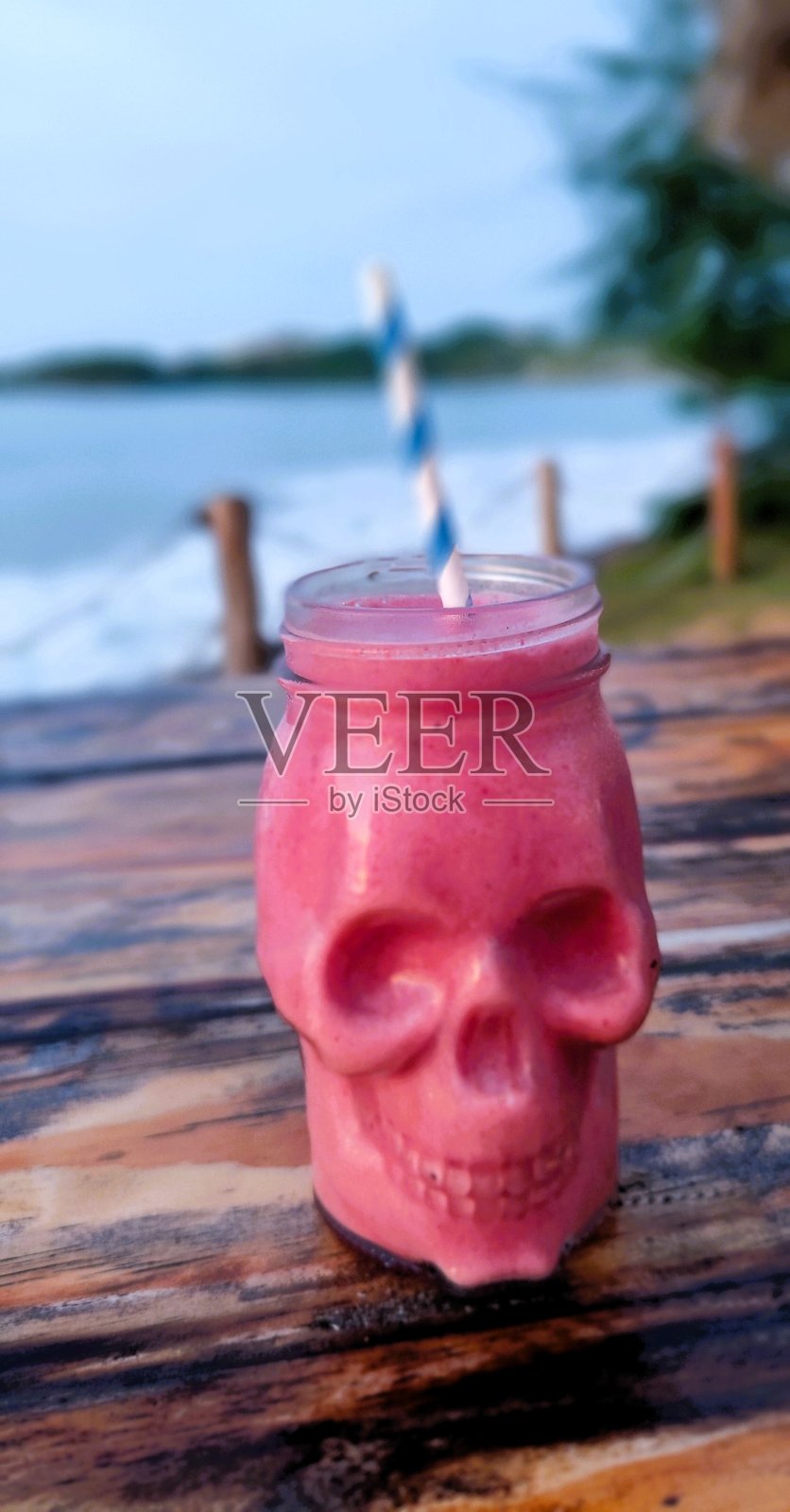 海滩上的骷髅形玻璃杯中装着热带粉色冰沙，配有吸管照片摄影图片