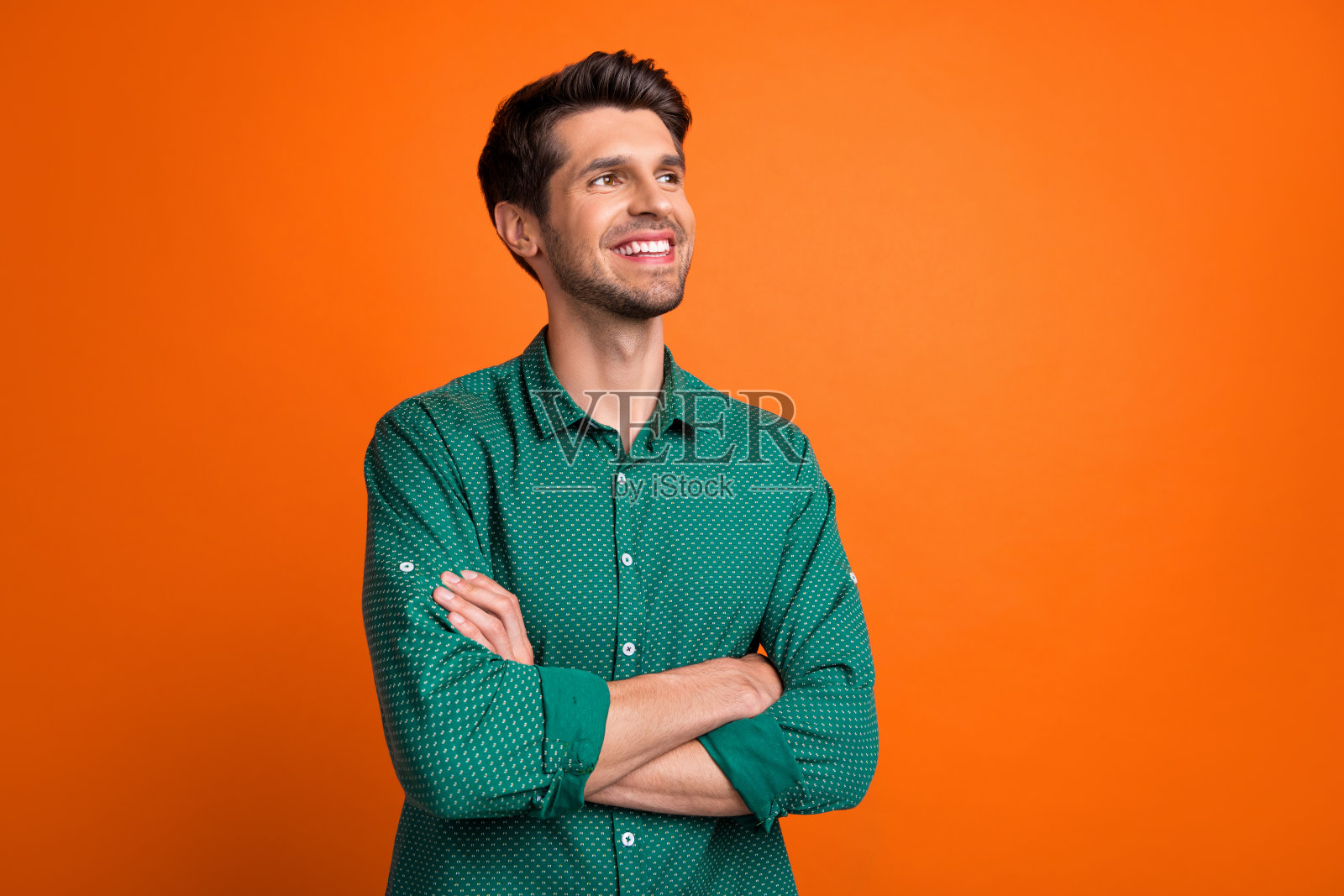 穿着绿色衬衫、双臂交叉的自信梦幻男子，凝视空旷的空间，背景为橙色。照片摄影图片