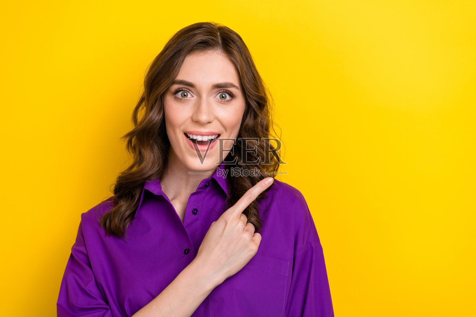 惊讶的女孩肖像，卷发披肩，穿着紫色上衣，指向空白的促销空间，目光注视着黄色背景。照片摄影图片