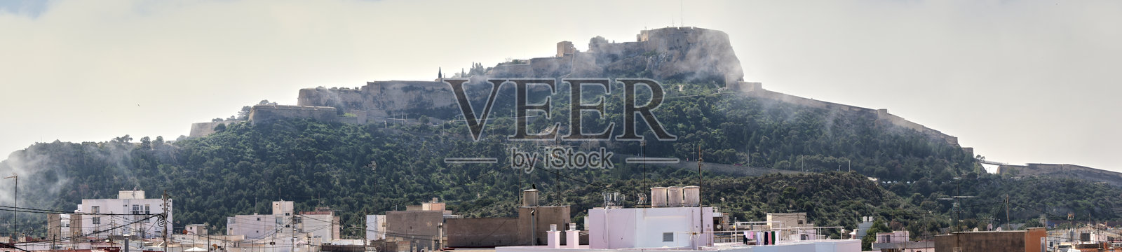 雾气弥漫的阿利坎特城堡全景，俯瞰城市屋顶和郁郁葱葱的绿地照片摄影图片