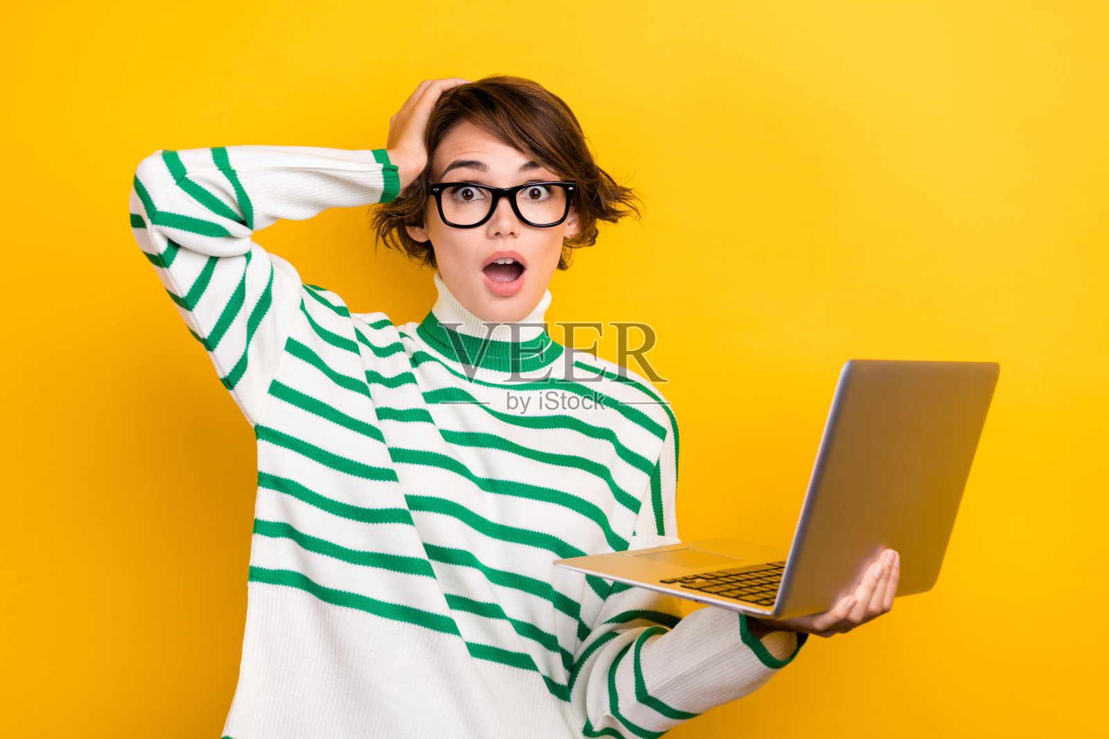 一位惊讶的无言女士，手持笔记本电脑，手触头部，张嘴凝视，背景为黄色。照片摄影图片