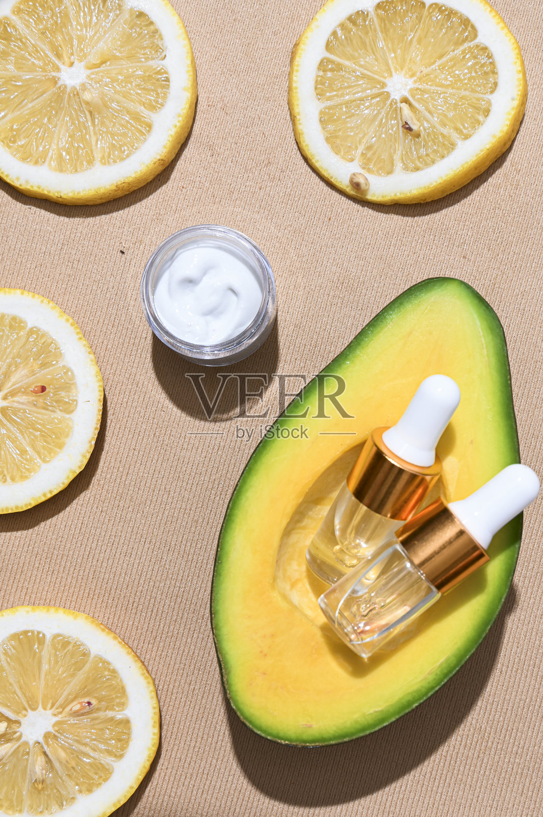 两个透明的护肤滴管瓶，配有金色瓶盖，放置在新鲜的鳄梨半边，周围环绕着绿色叶子，背景为质感米色。自然美和有机护肤概念。照片摄影图片