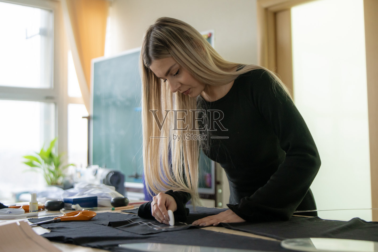 一位纺织艺术家在明亮的工作室里小心翼翼地用粉笔在黑色布料上画线，周围摆放着各种缝纫工具和材料。照片摄影图片