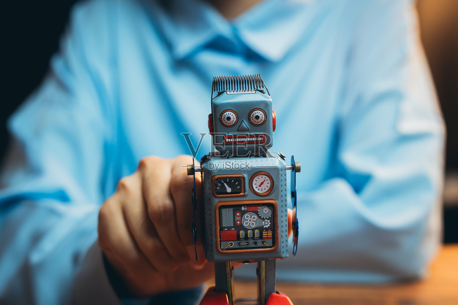穿着蓝色衬衫的人手持复古玩具机器人，放在木桌上，背景中性。照片摄影图片