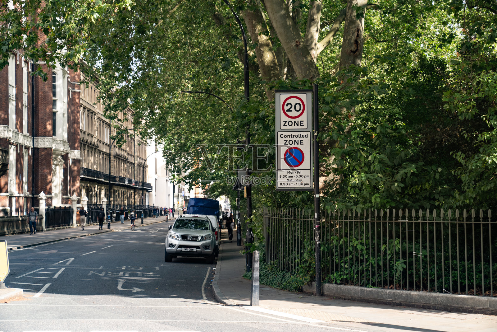 城市街道上有限速和管控区域标志，旁边停着汽车和树木成行的人行道，在阳光下。照片摄影图片