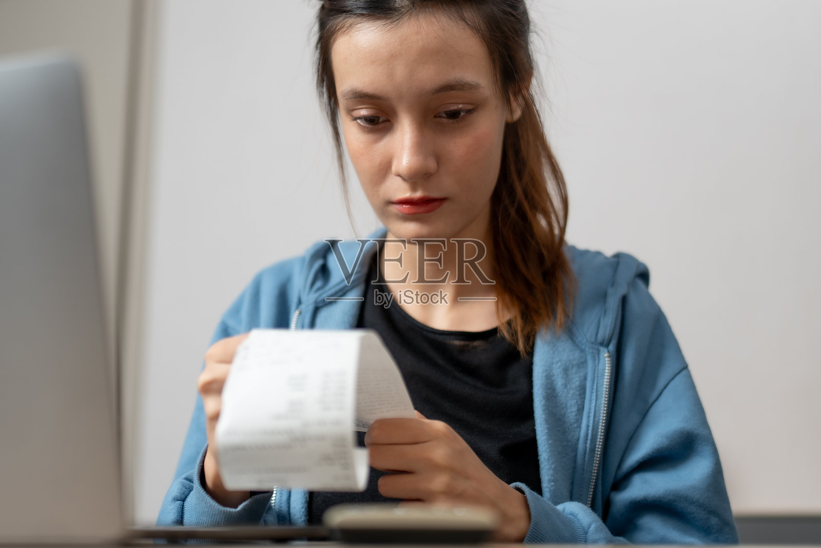 年轻女性坐在桌子前，专注而沉思地查看收据，旁边有一台笔记本电脑。她在思考自己的开支。照片摄影图片