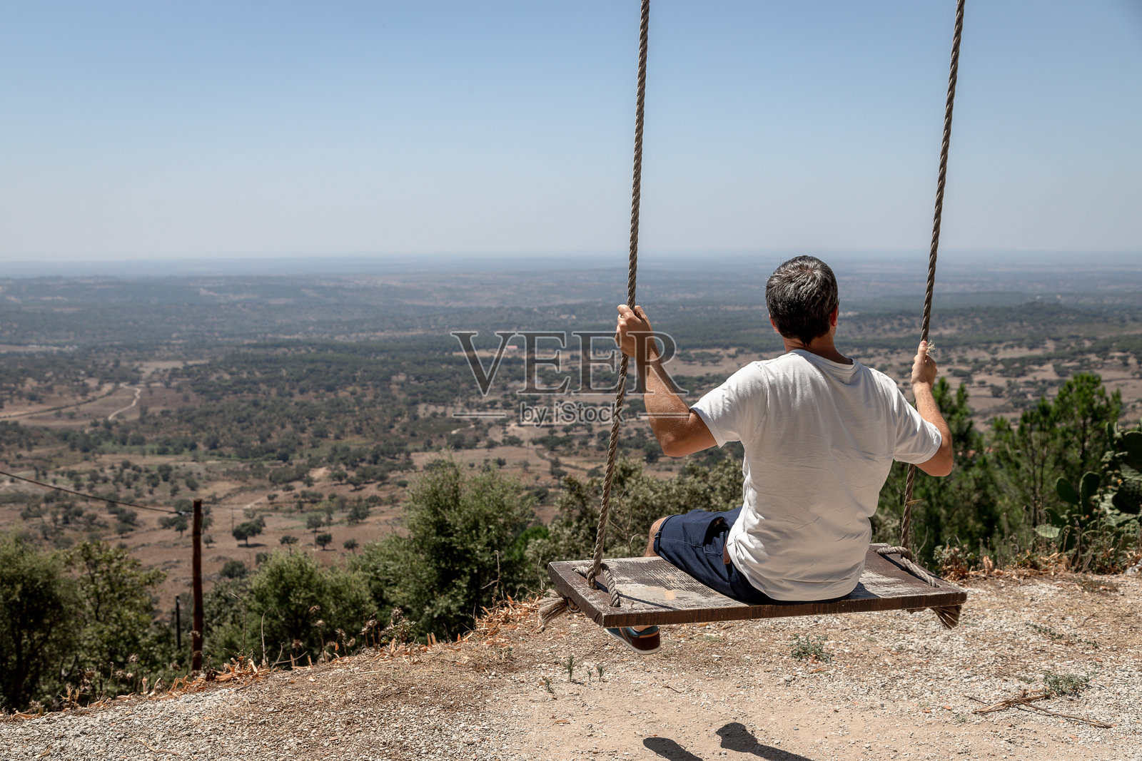 一名男子在葡萄牙阿连特茹地区波尔塔莱格雷的卡雷拉斯，福特卡尔沃埃罗斯观景点的木秋千上享受着宁静的时光，俯瞰着广阔的风景，炎热的夏日里。照片摄影图片