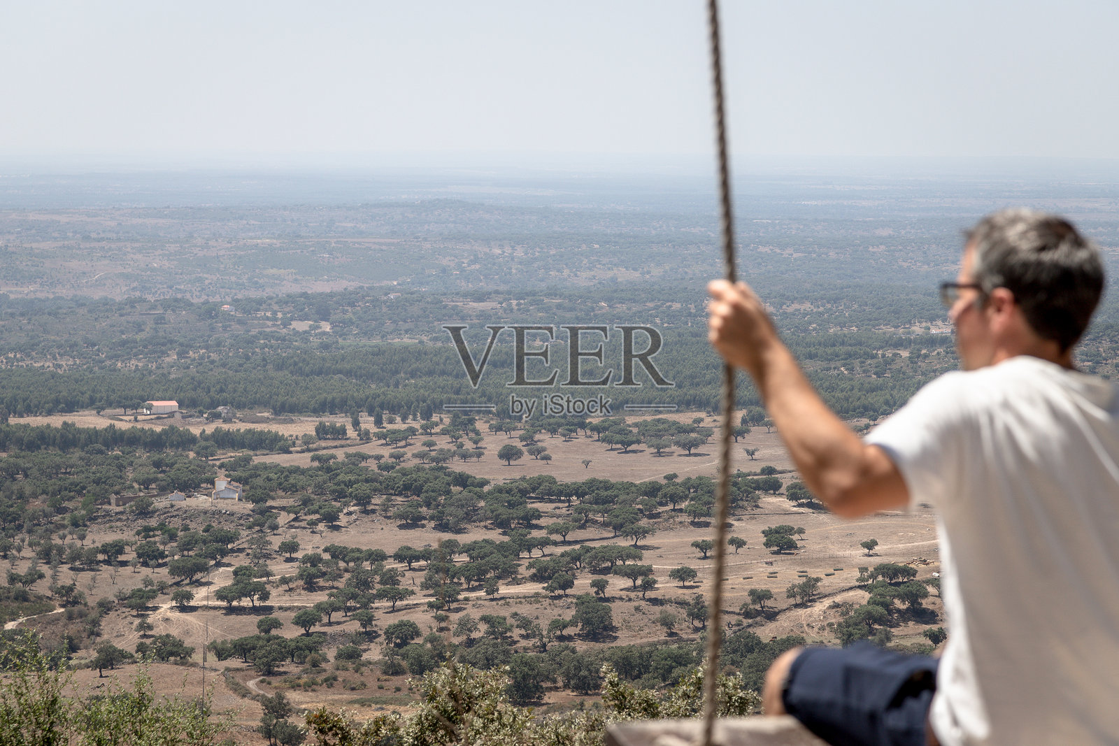 一名男子在葡萄牙阿连特茹地区波尔塔莱格雷的卡雷拉斯，福恩特·多斯·卡尔沃埃罗斯观景点的木秋千上享受着宁静的时光，俯瞰着广阔的风景，炎热的夏日里。照片摄影图片