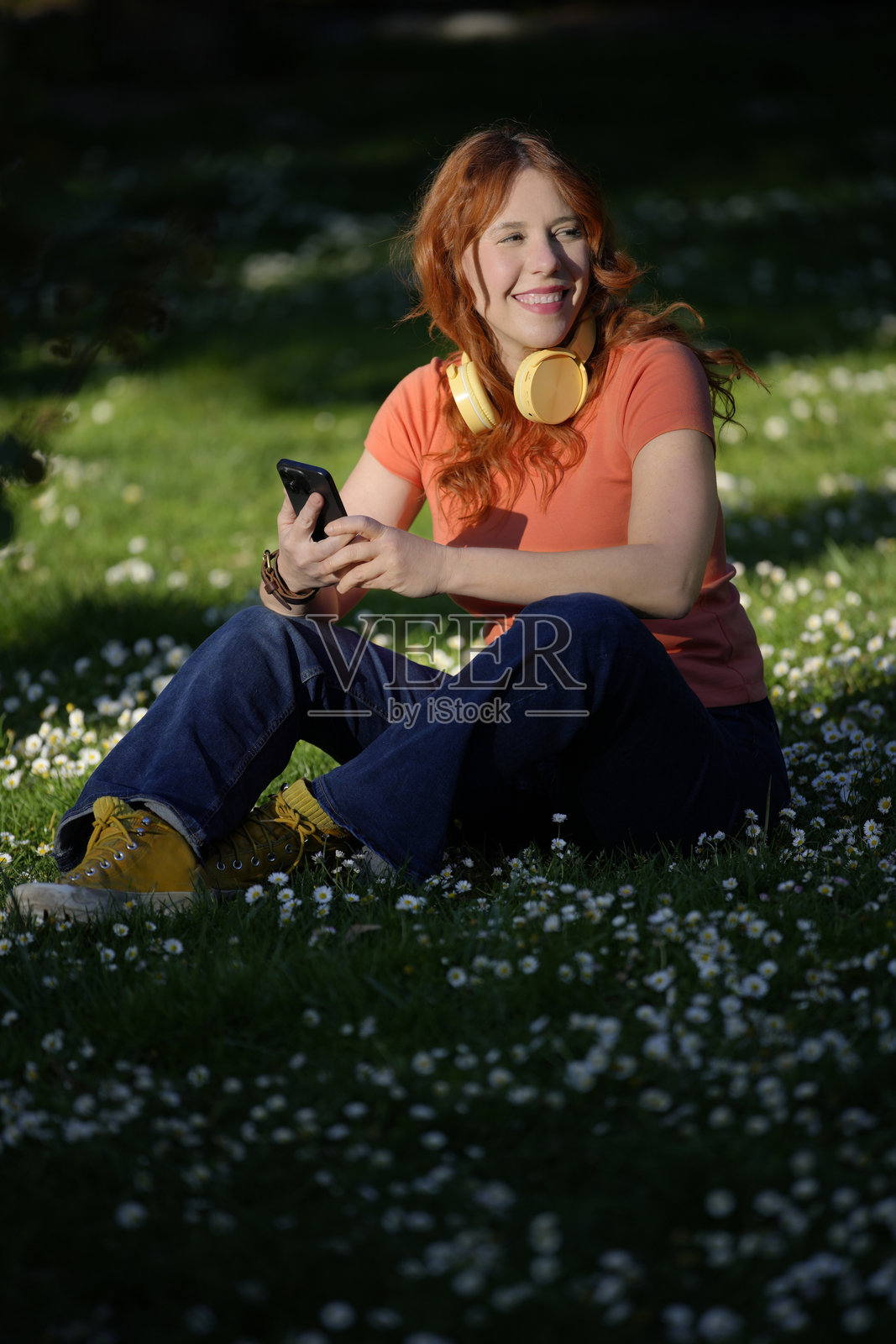 年轻女性坐在草地上，戴着耳机，微笑着使用智能手机。照片摄影图片