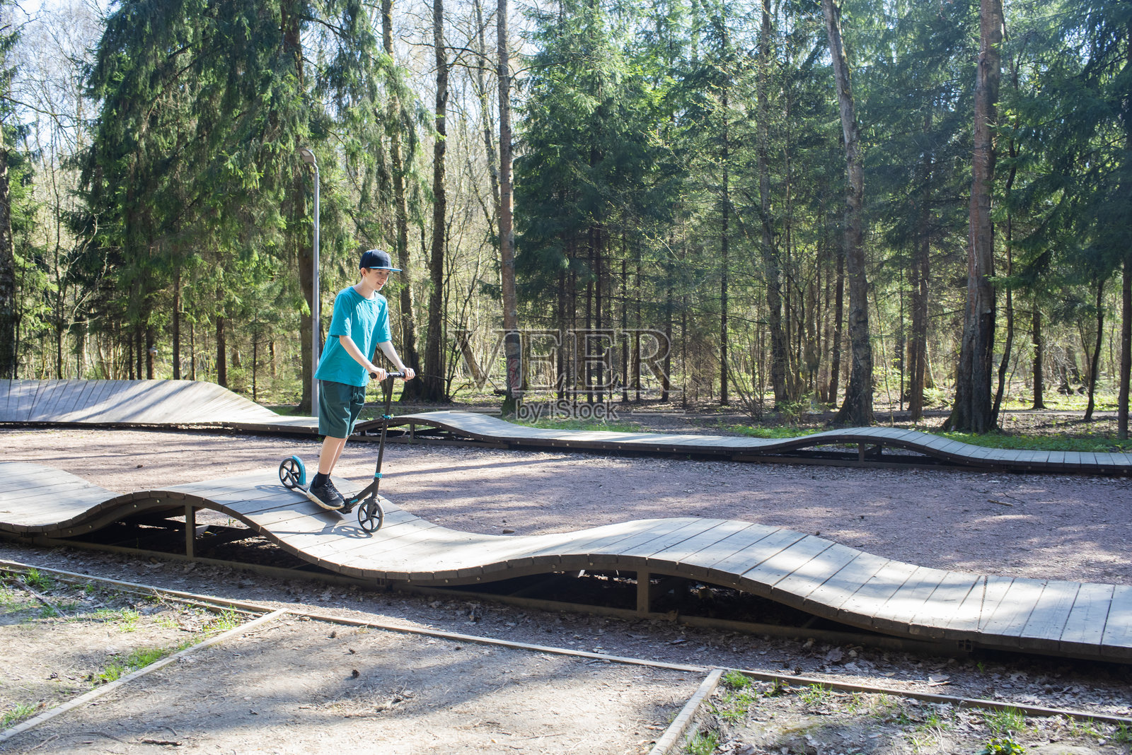 一个12岁的小男孩戴着帽子和短裤，在莫斯科的博罗夫斯基公园的泵道上骑滑板车，春天的景象。照片摄影图片