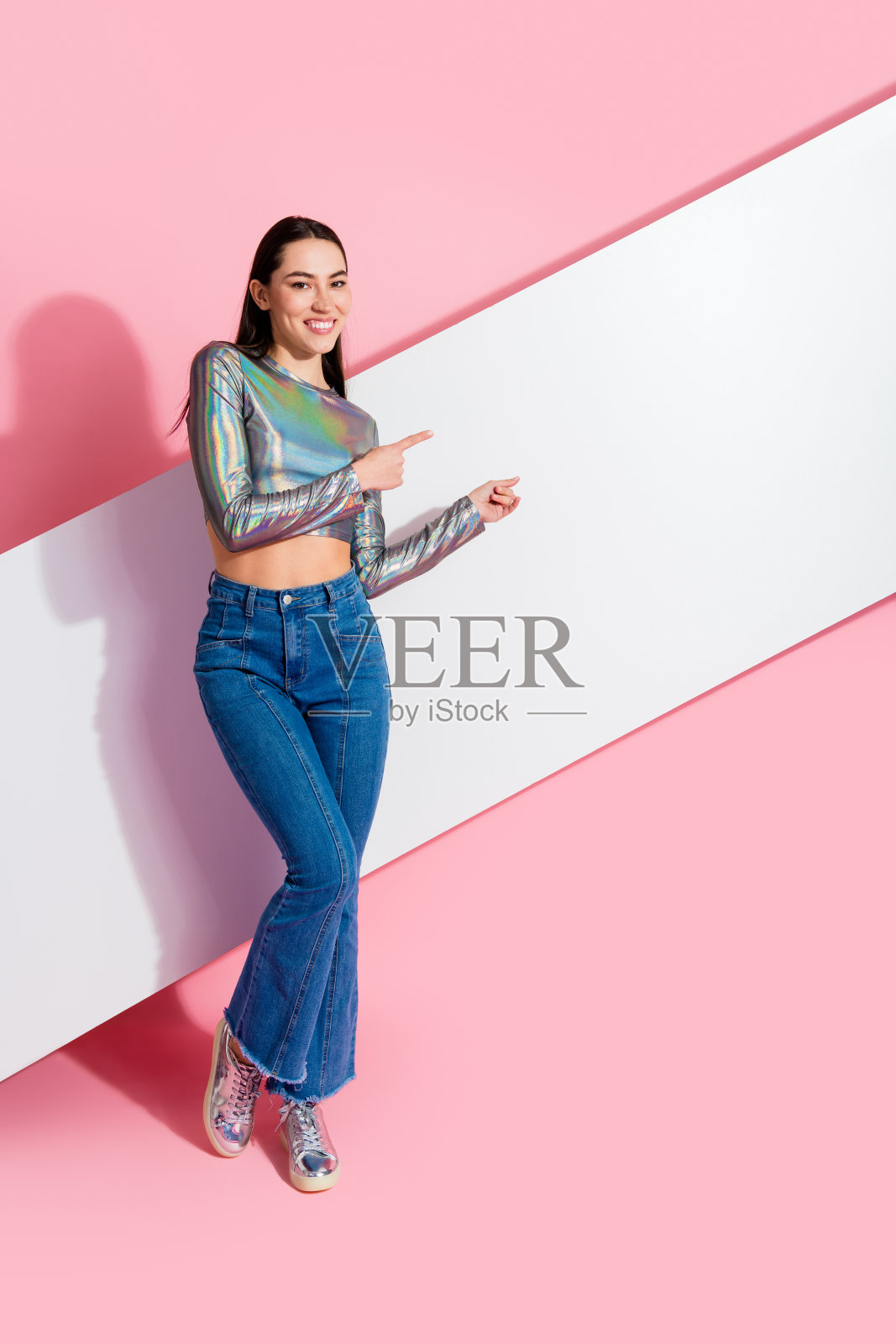 年轻时尚女性自信地微笑，指向粉色竖墙上的空白处，展现出休闲而迷人的时尚风格。照片摄影图片