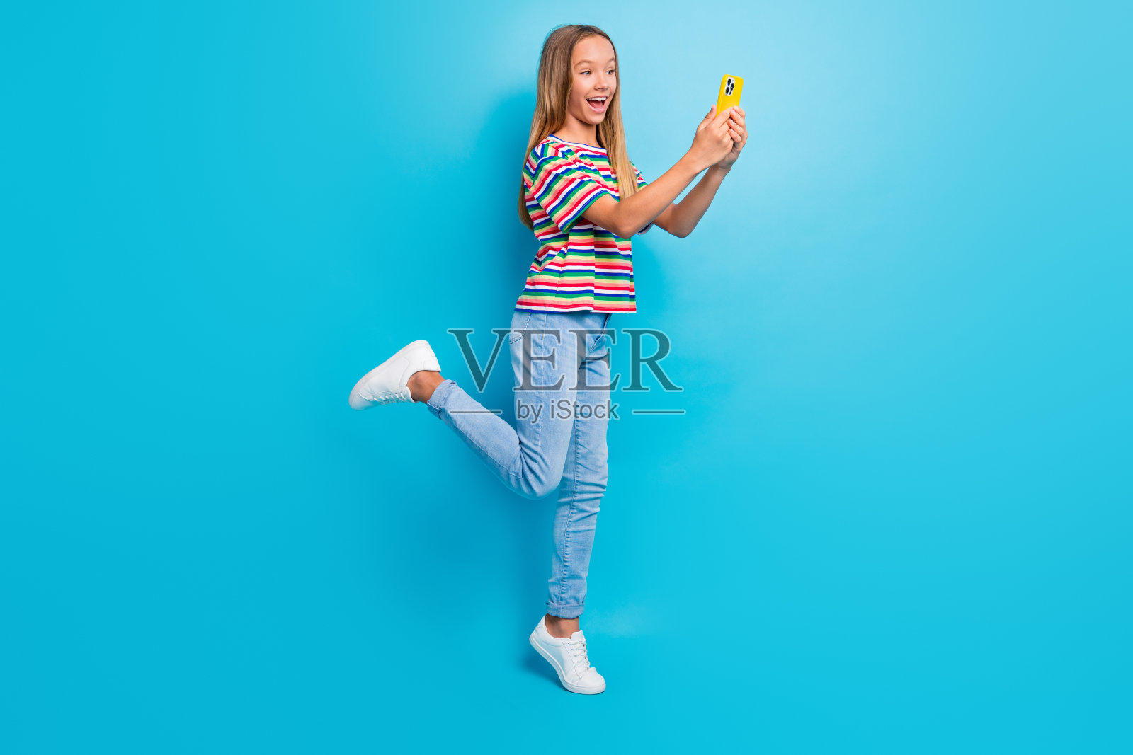 迷人的少女穿着彩色T恤和牛仔裤，在蓝色背景下用智能手机自拍的全身照片照片摄影图片
