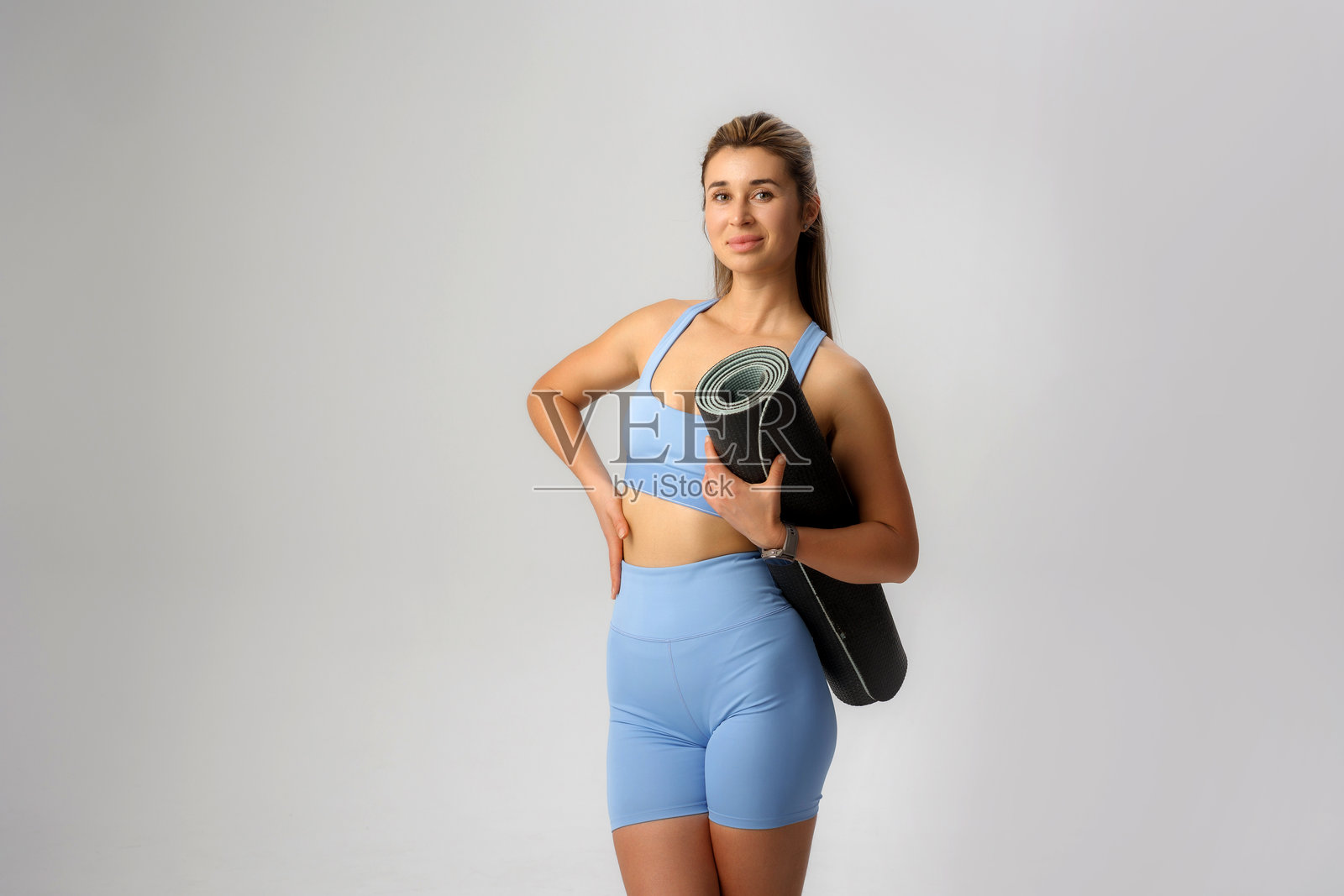 瑜伽练习者自信地摆姿势，手持瑜伽垫，展示健身与健康的生活方式，背景为一个瑜伽工作室。照片摄影图片