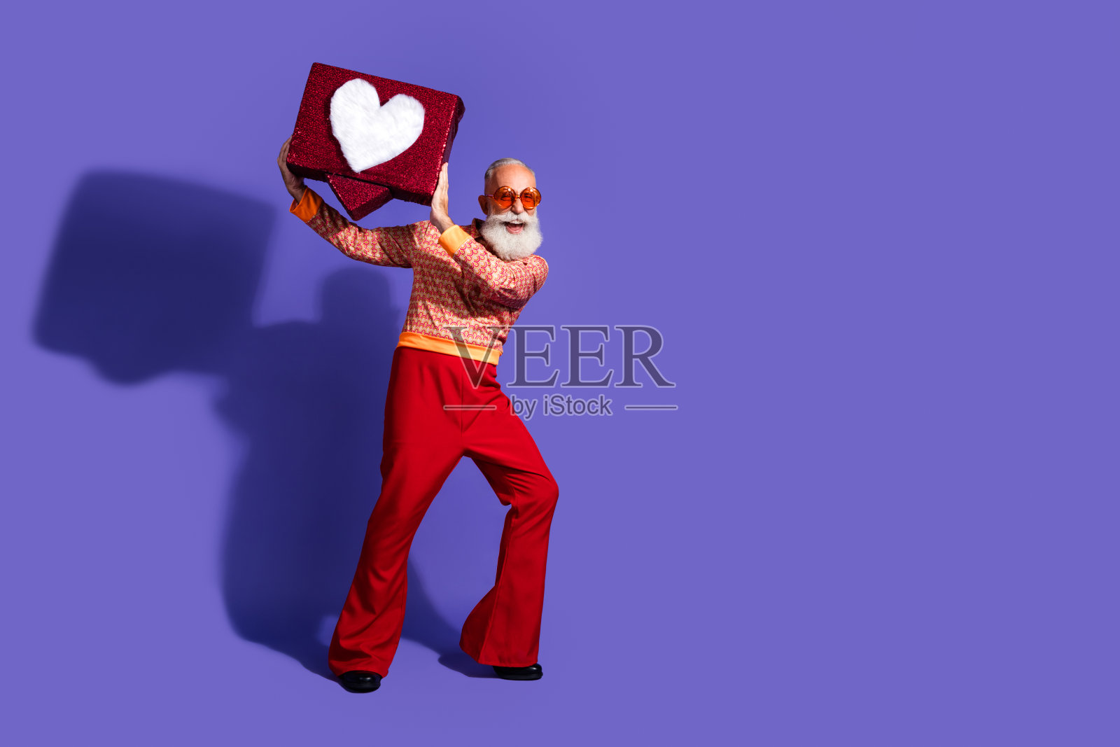 快乐的老年人手持心形礼品盒，背景为鲜艳的紫色，展现复古风格的时尚。照片摄影图片