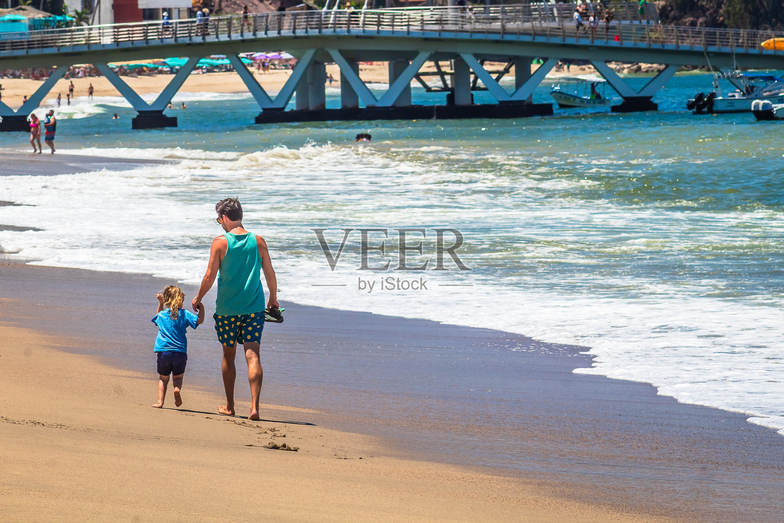 一位父亲与他的小女儿在海滩上散步，背景是哈利斯科州普埃尔托·瓦亚尔塔的建筑。照片摄影图片