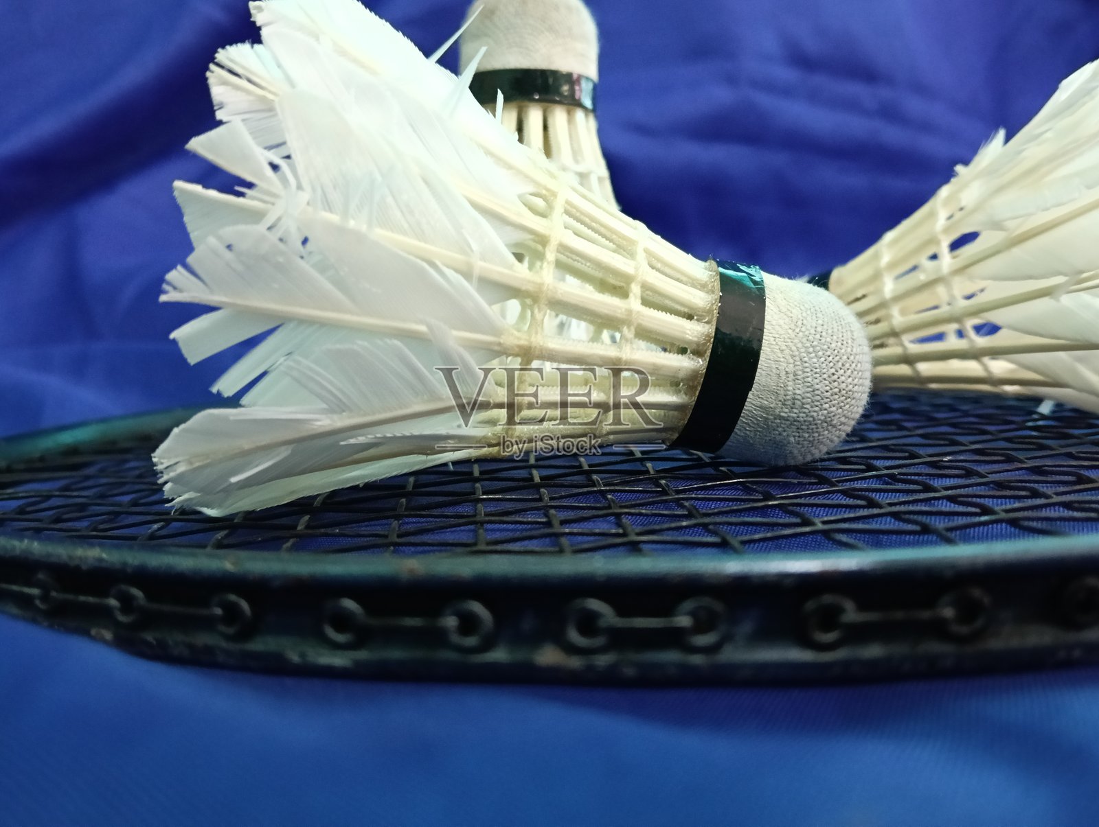 黑色羽毛球拍上放着磨损或使用过的羽毛球，背景为蓝色，特写。照片摄影图片