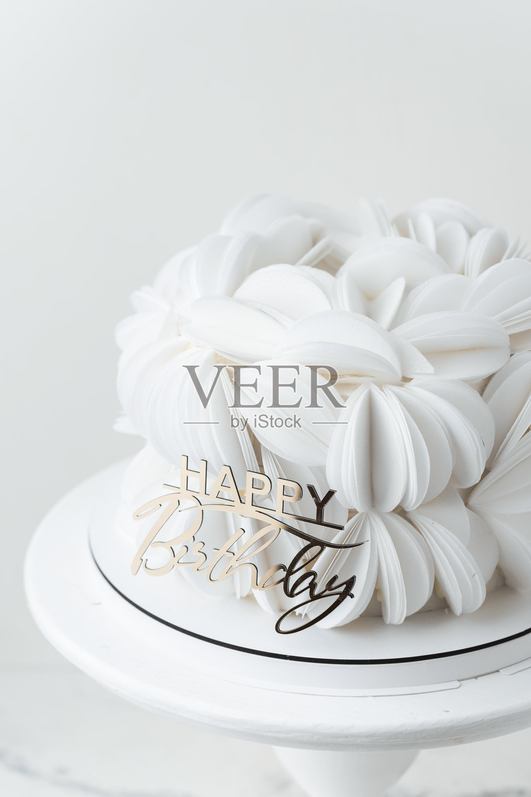 生日蛋糕上覆盖着白色可食用纸花瓣，装饰着金色的“生日快乐”顶部装饰。白色背景。照片摄影图片