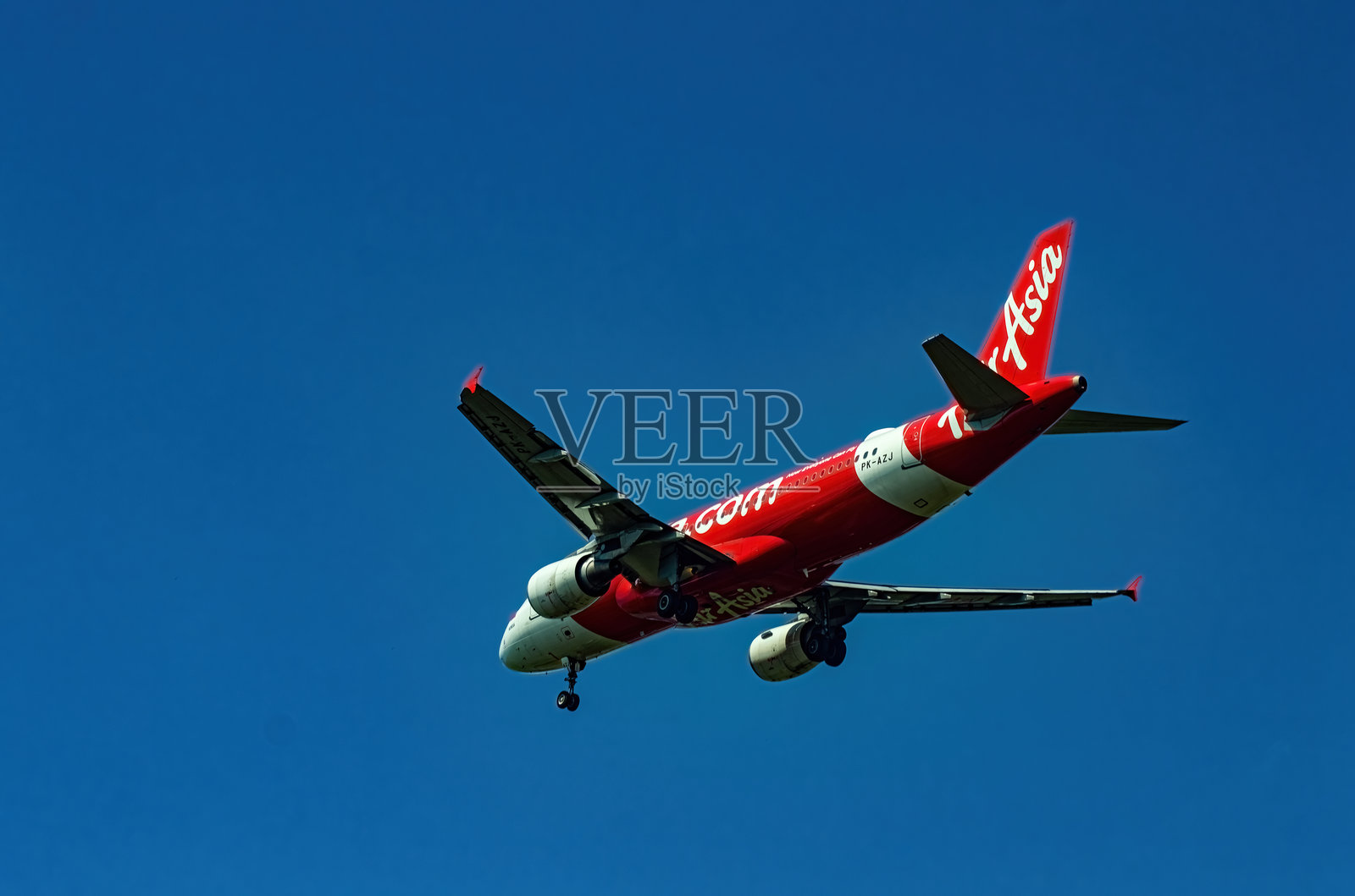一架涂有红白相间涂装的亚航飞机在明亮的蓝天中翱翔。飞机的引擎和机身在阳光下闪耀,保持着高度。印尼,2025年5月10日照片摄影图片