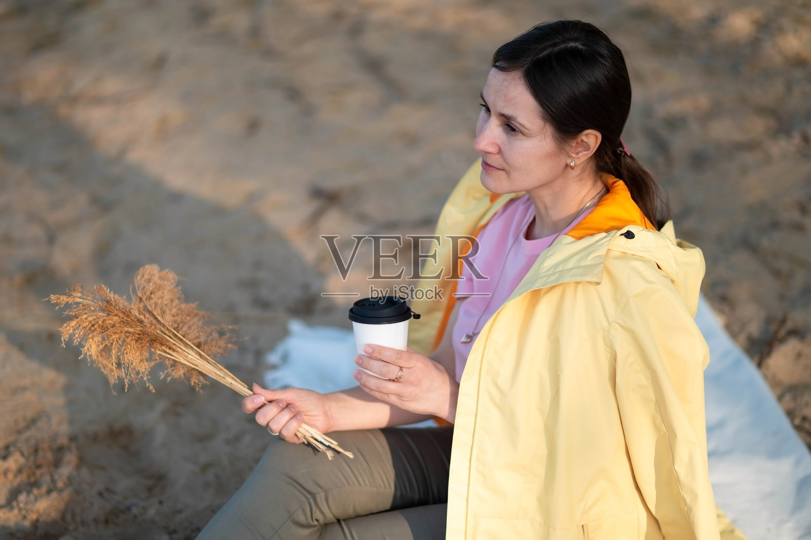 一位女性坐在湖边，手握纸杯喝热饮，手中还捧着一束干花。照片摄影图片