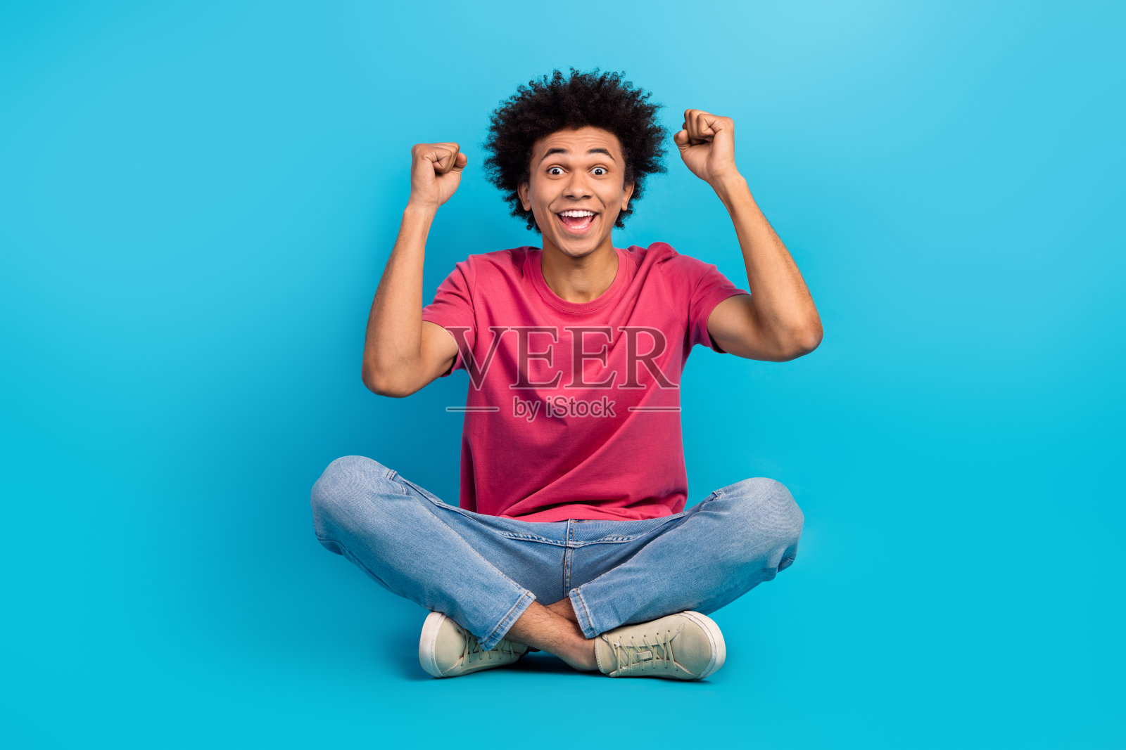 全身照：一位吸引人的年轻男子兴奋地坐在地板上，双拳举起，穿着时尚的粉色衣服，背景为蓝色。照片摄影图片