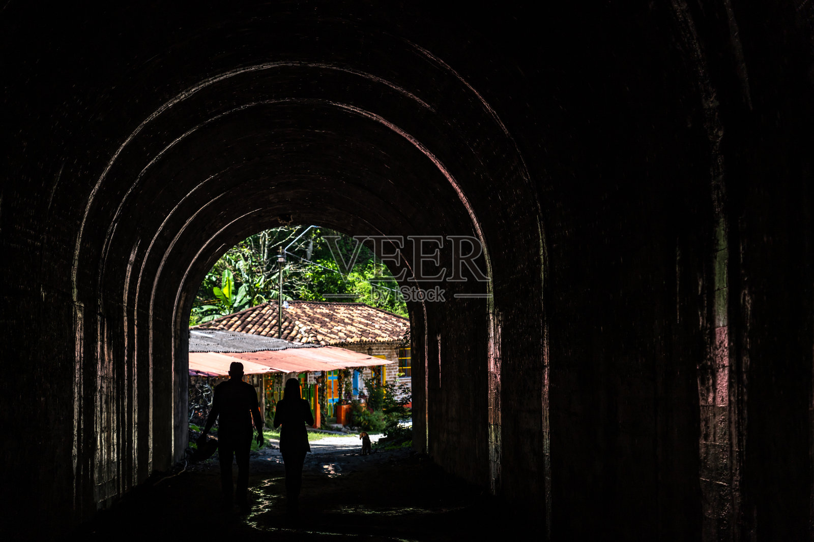 两个身影在一个废弃潮湿的隧道里行走。黑暗中的冒险与发现，背景是一座传统房屋。照片摄影图片