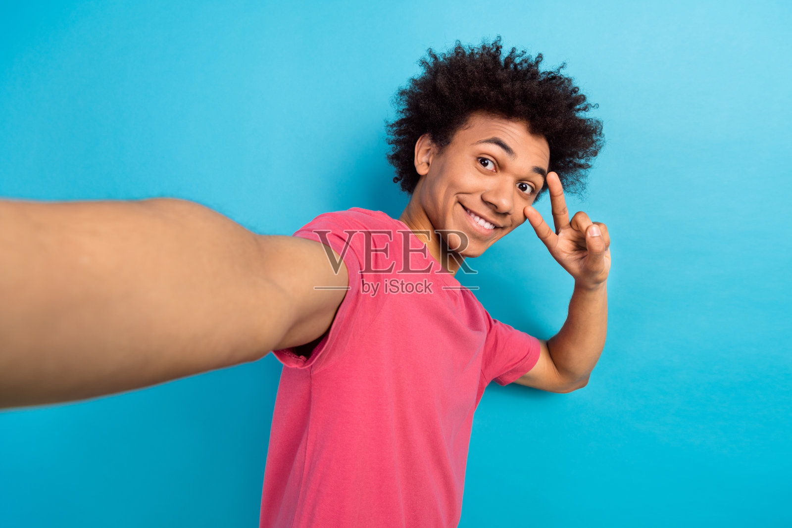 乐观的好男人穿着粉色T恤，做着自拍，眼睛上做出V字手势，背景为蓝色。照片摄影图片