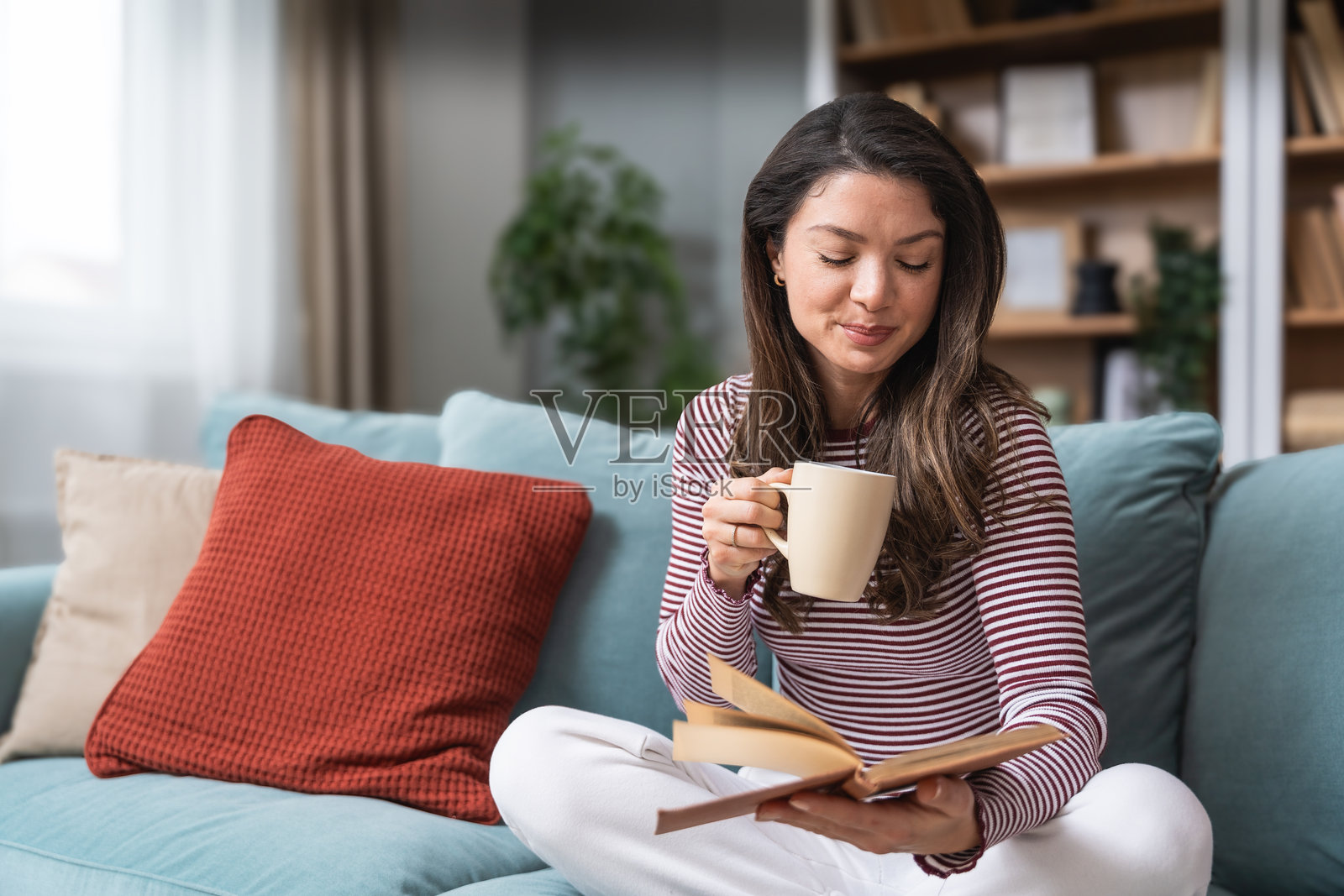 生活平衡、自尊心、心理健康和独立性。一位年轻女性坐在她的小型现代家中，喝着咖啡，阅读一本书。她保持心理稳定并自我教育。照片摄影图片