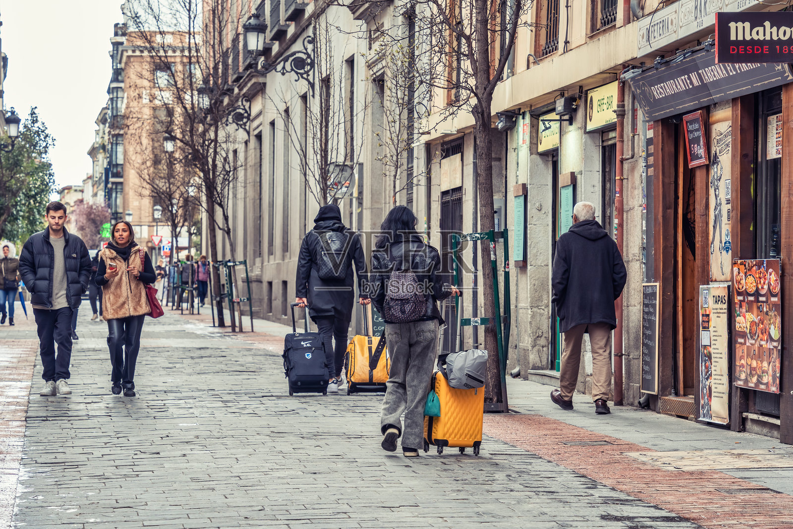 一群旅行者在西班牙马德里的鹅卵石街道上行走，拖着他们的行李箱。照片摄影图片