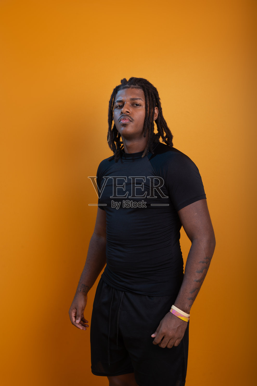 穿着短裤和黑色衬衫的帅气男子，扎着辫子，站在橙色背景前摆姿势拍照。照片摄影图片