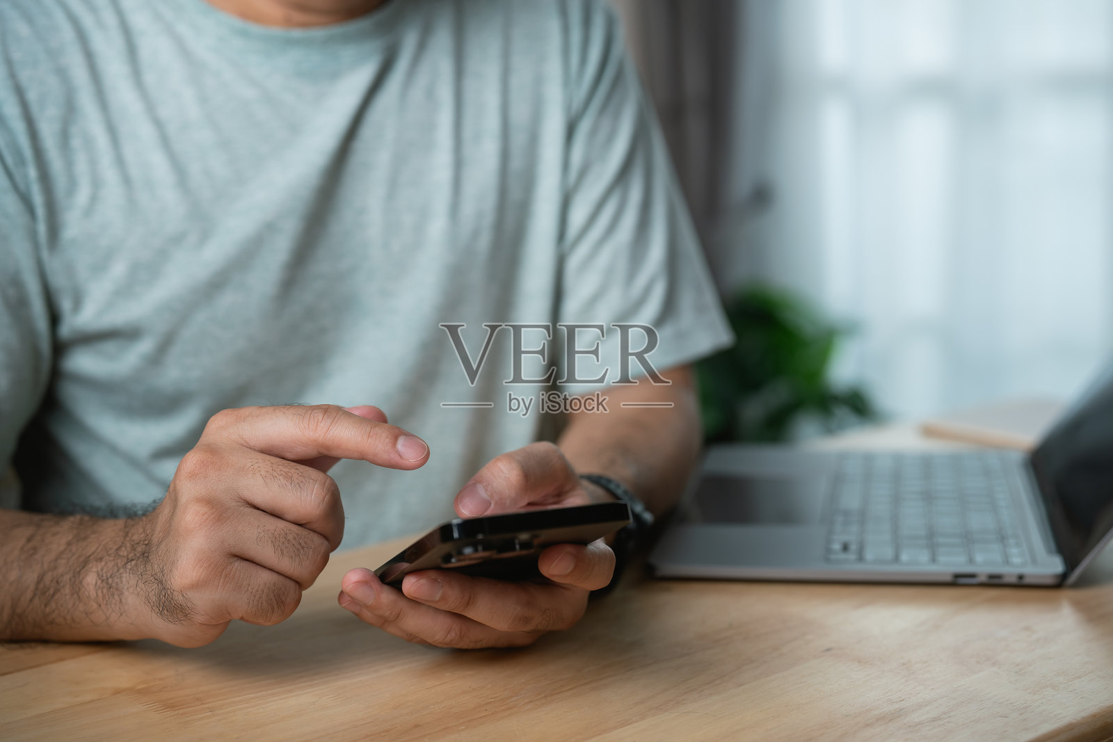 在现代家居办公室环境中，坐在木桌旁使用智能手机的人，旁边有一台笔记本电脑，自然光透过窗户洒入。照片摄影图片